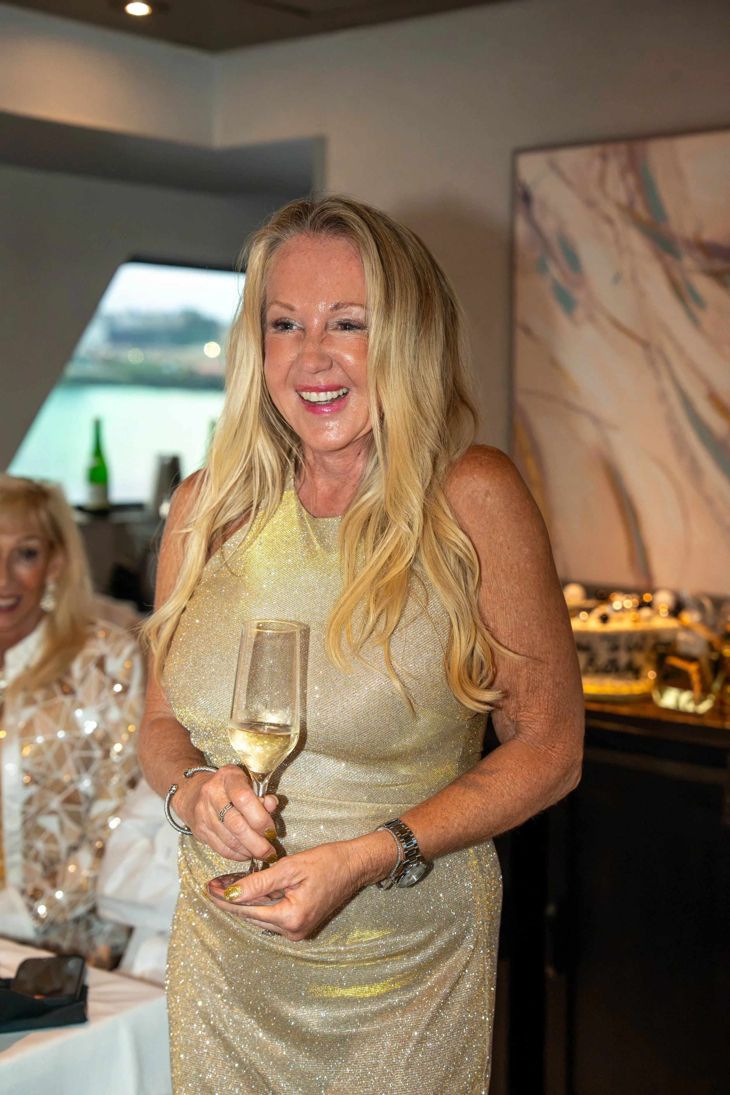 Smiling blonde woman in a sparkling gold dress holding a wine glass at an indoor celebration.