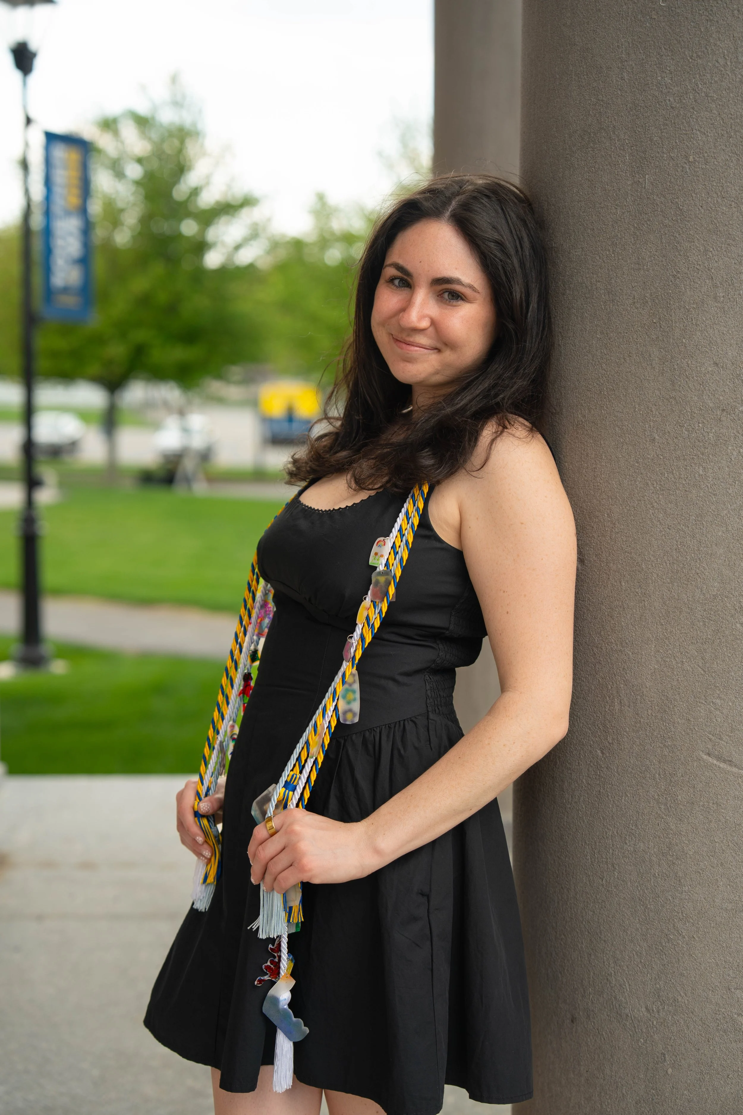 Smiling woman in a black dress wearing colorful graduation honor cords outdoors
