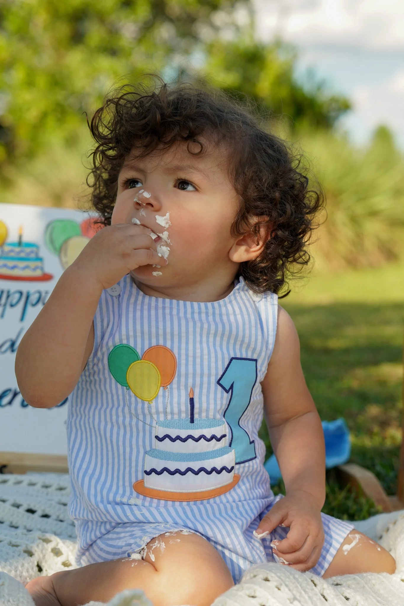 Child eating birthday cake outdoors while wearing a blue striped outfit with a birthday cake design