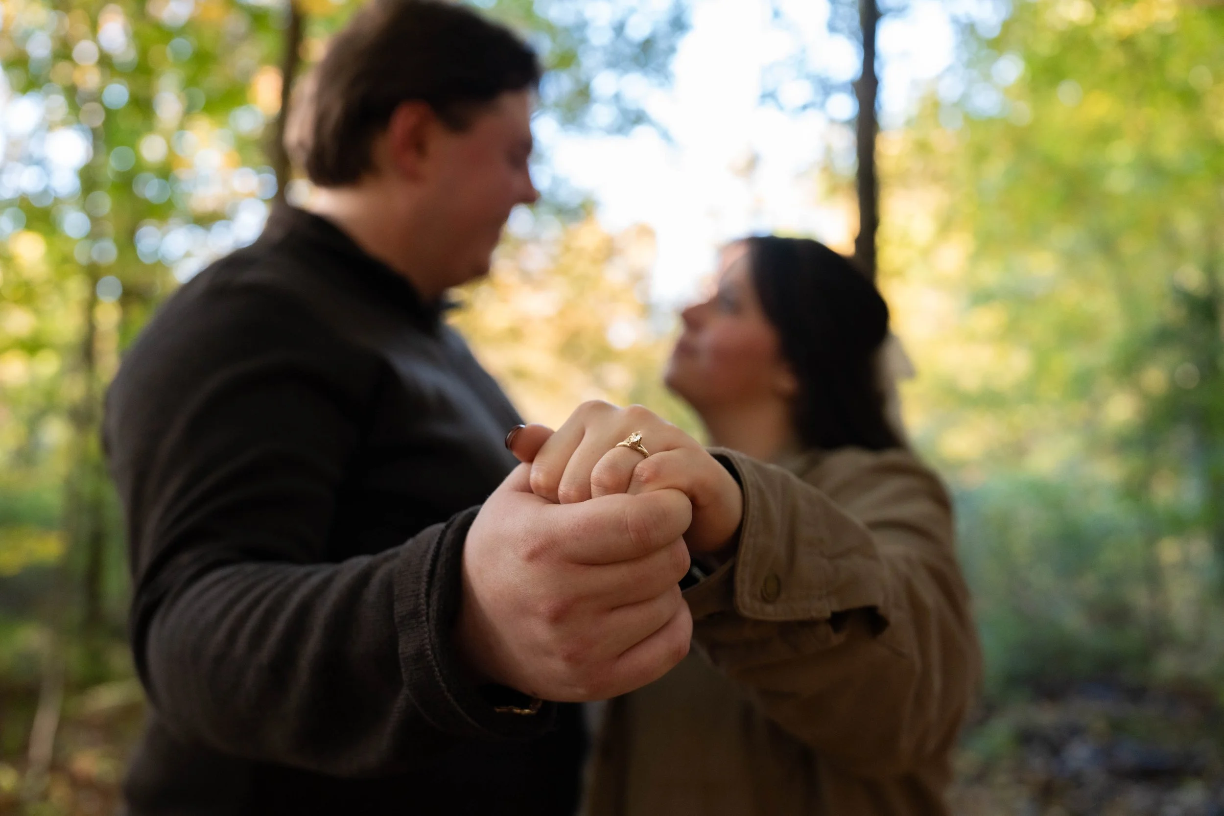 Couple holding hands and showing an engagement ring outdoors in a sunlit forest