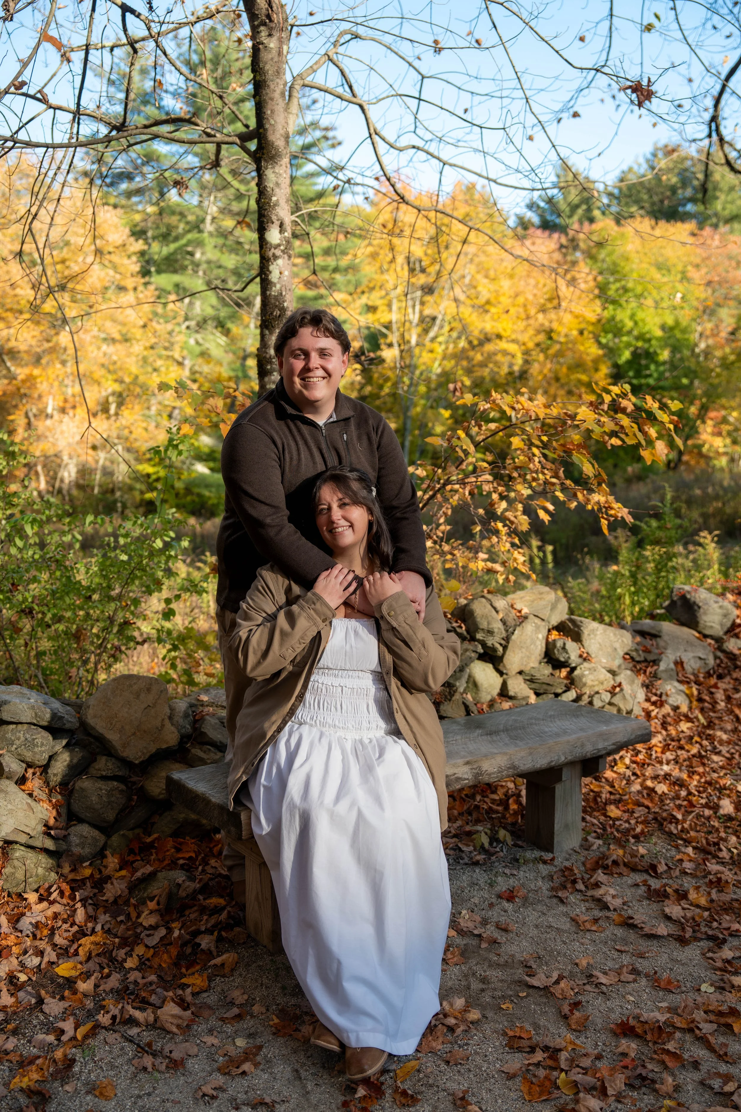 Two smiling people posing together in an autumn forest, with one standing behind the other on a wooden bench.