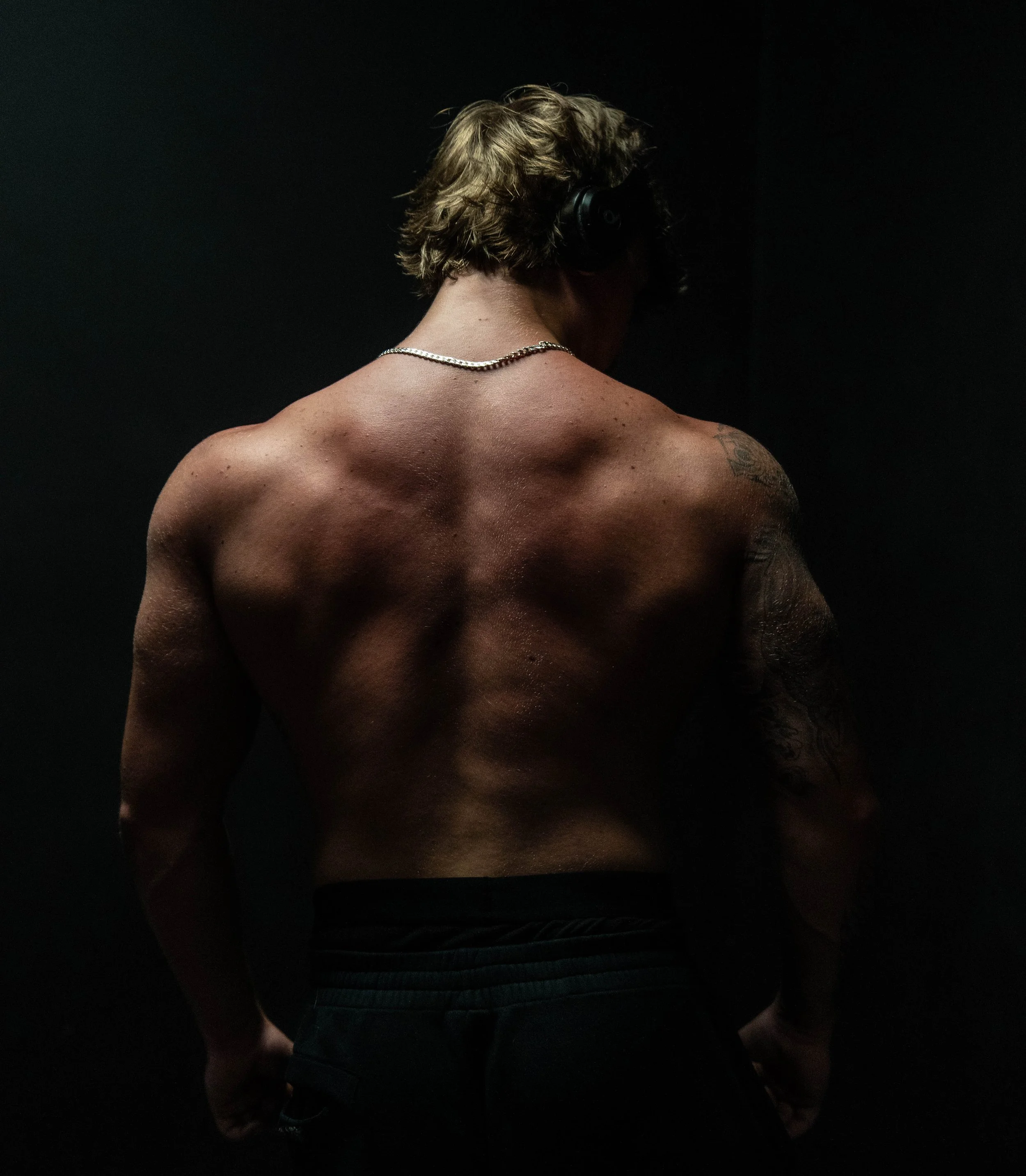 Muscular shirtless man seen from behind wearing headphones and a chain necklace against a black background