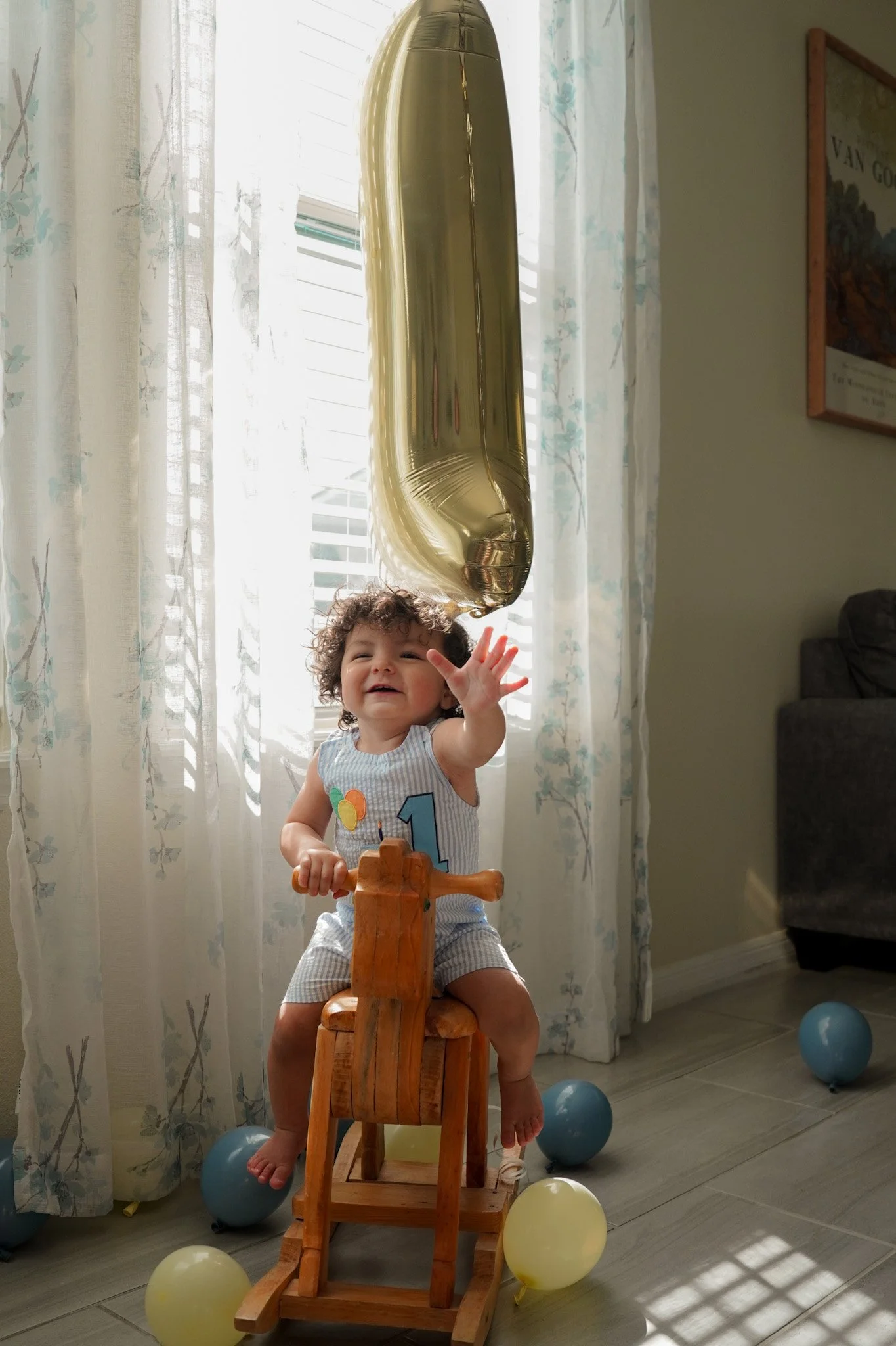 Smiling baby in a number-one outfit riding a wooden rocking horse beneath a large gold balloon, surrounded by colorful balloons.