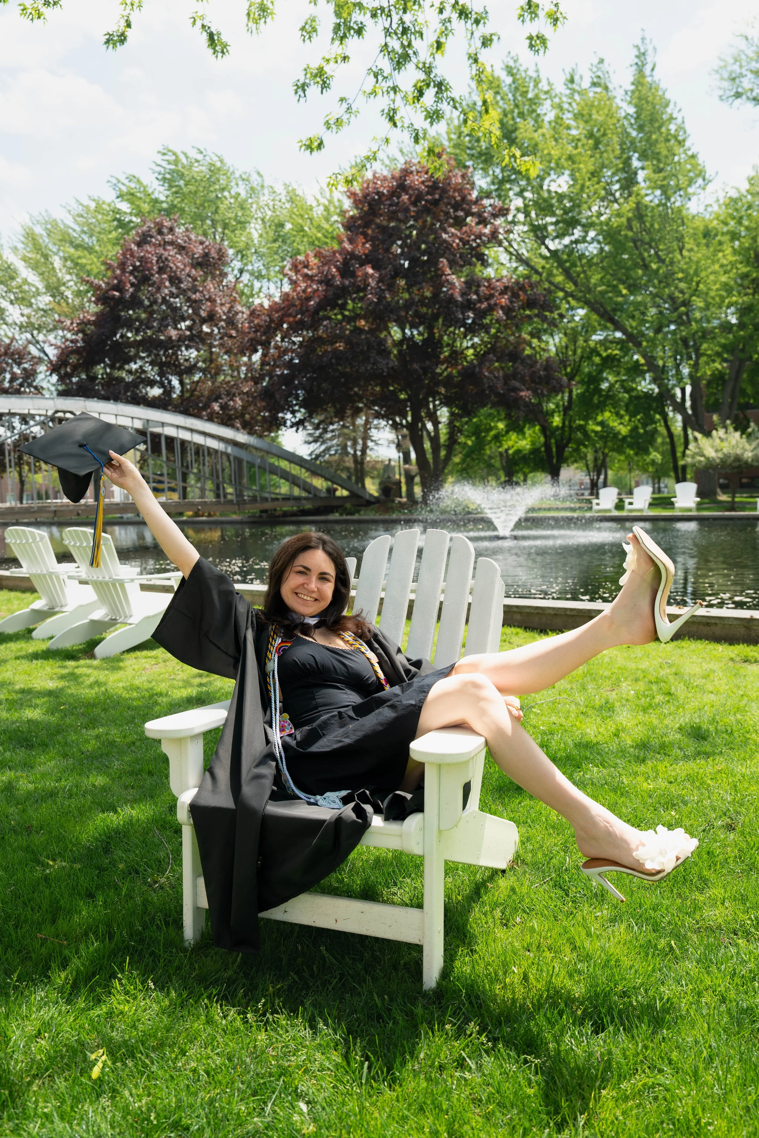 Smiling graduate in a black cap and gown relaxing in a white Adirondack chair beside a fountain and pond