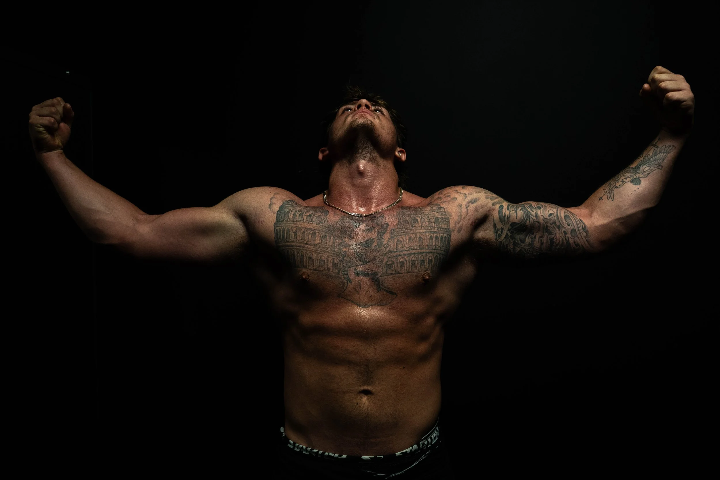 Muscular shirtless man with tattoos flexing both arms against a dark background