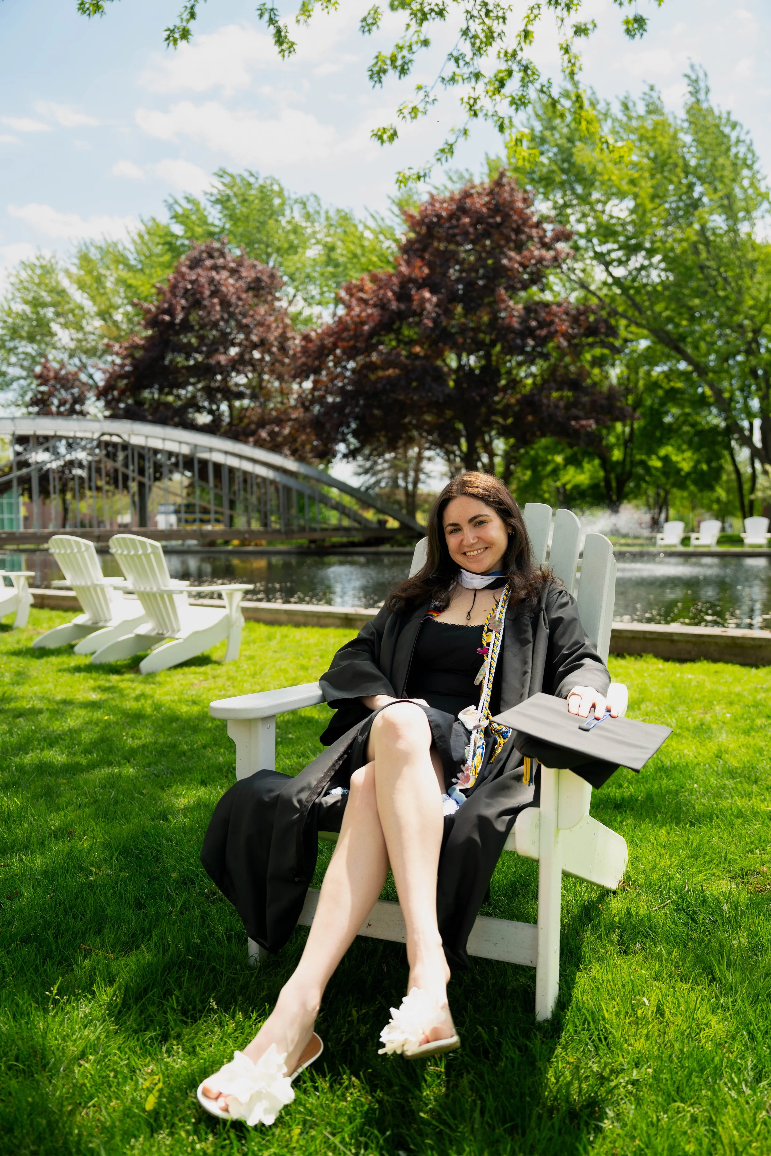 Smiling woman in a graduation cap and gown sitting in a white Adirondack chair beside a pond and arched bridge in a sunny park