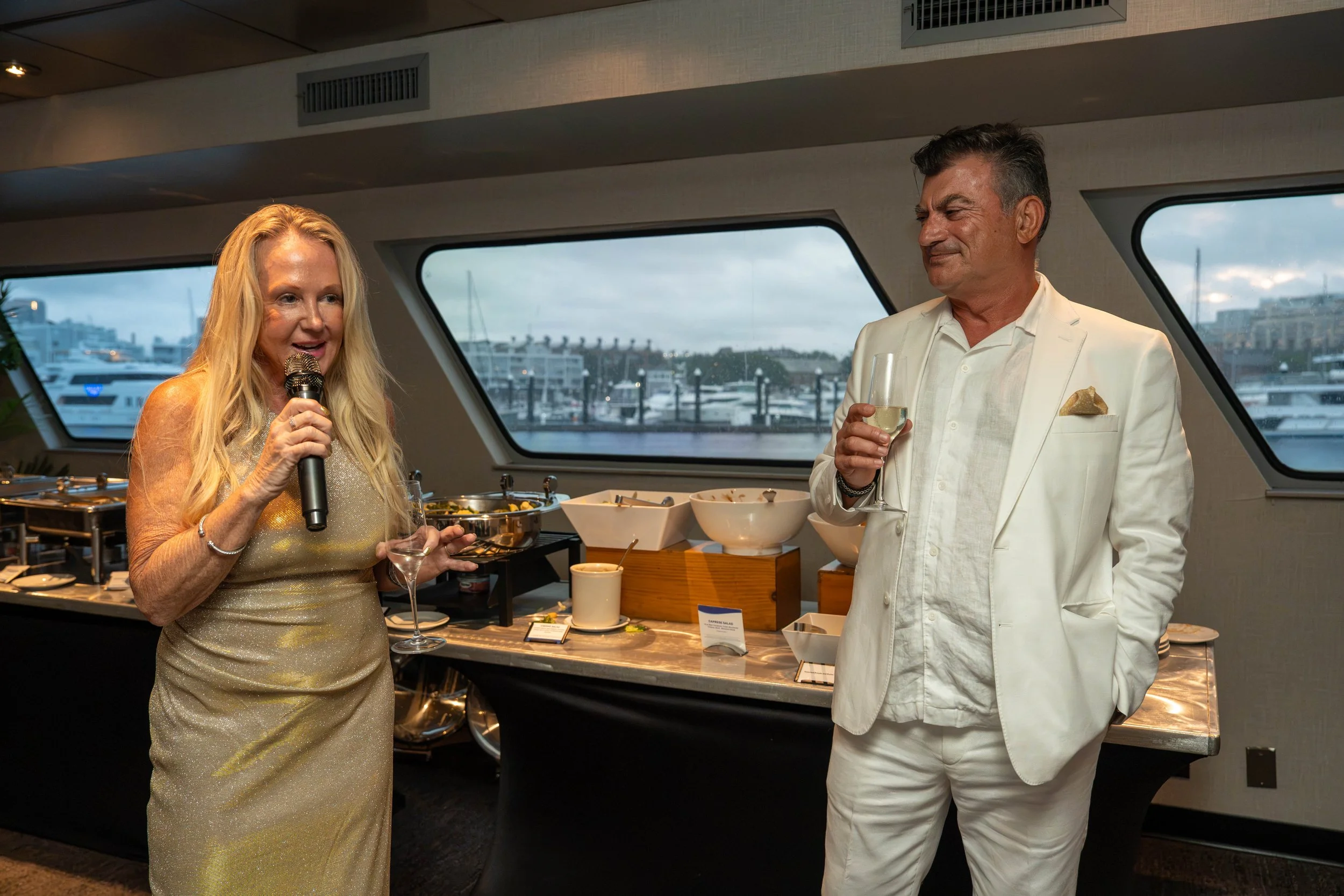 Woman speaking into a microphone beside a man in a white suit at a yacht buffet overlooking a marina.