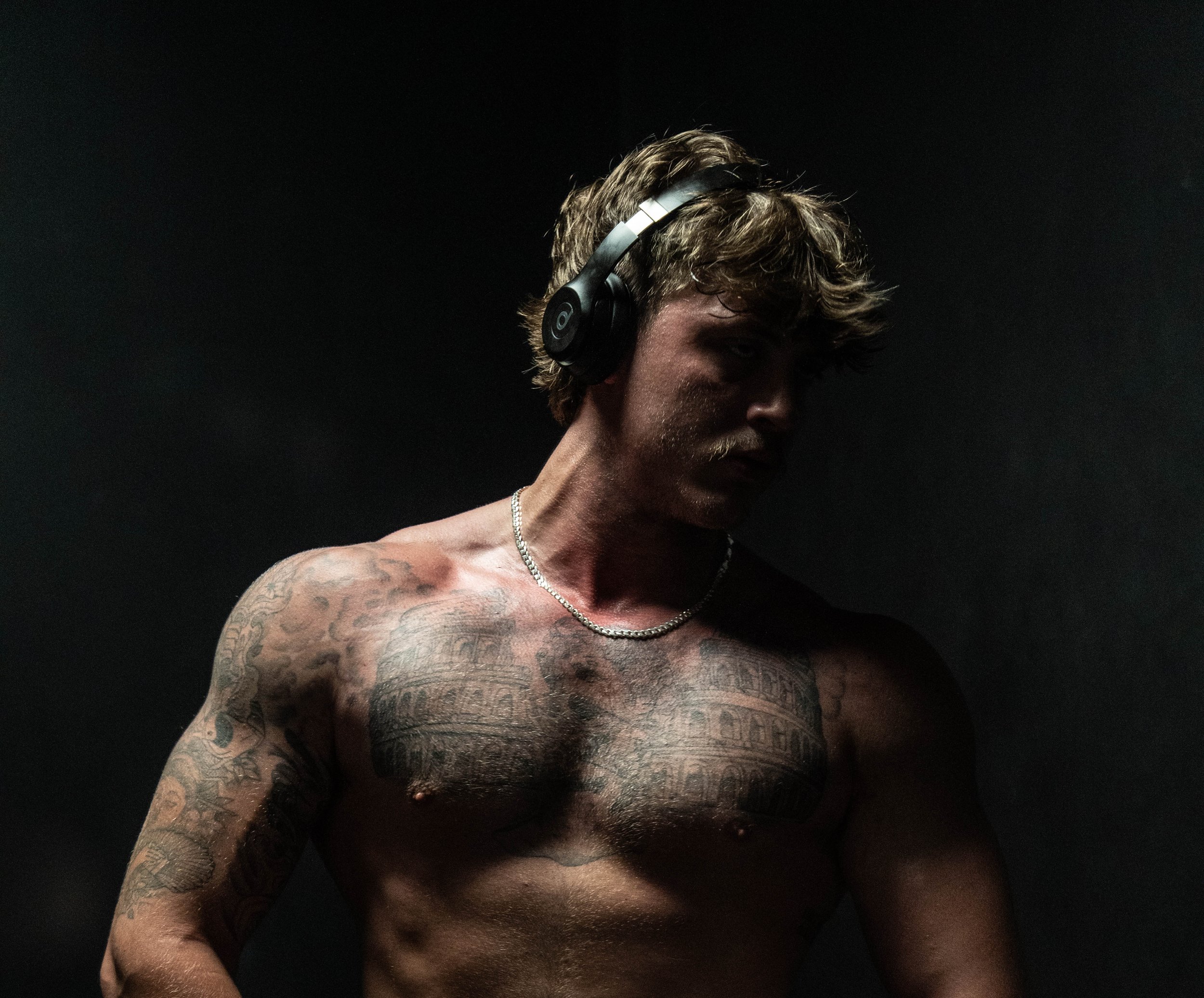 Tattooed shirtless man wearing black over-ear headphones in dramatic low-light portrait