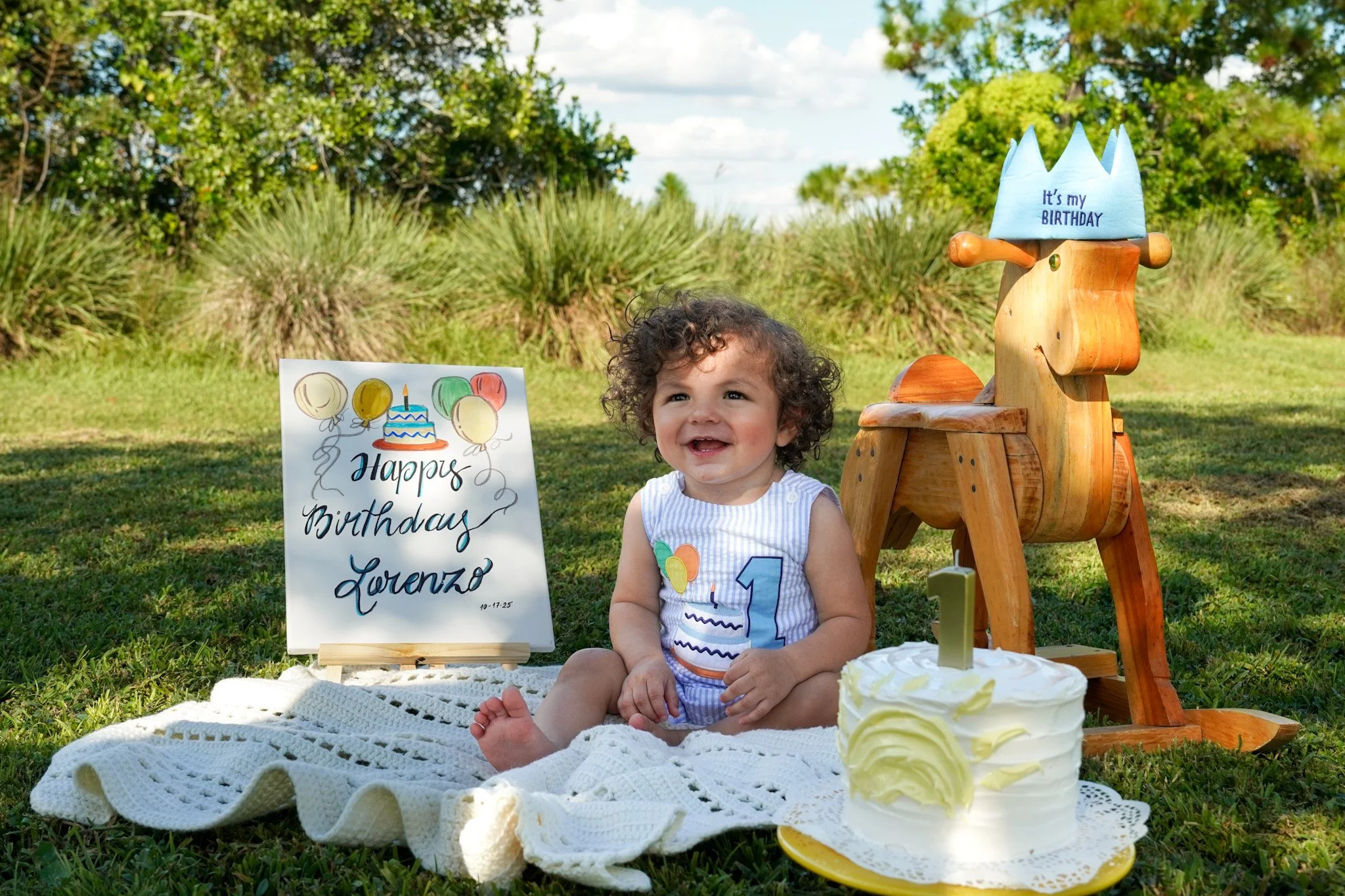 Smiling toddler celebrating a birthday outdoors beside a decorated cake, birthday sign, and wooden rocking horse wearing a party hat