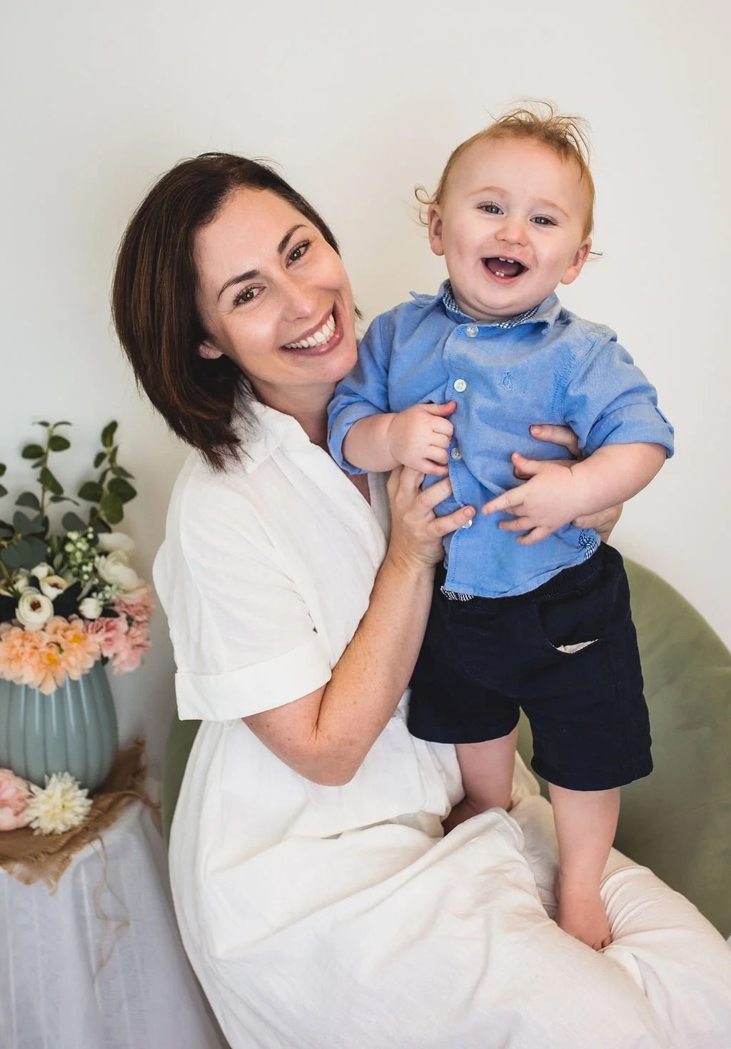 Mother holding her baby and both are smiling happily together seated on a chair with some flowers next to them