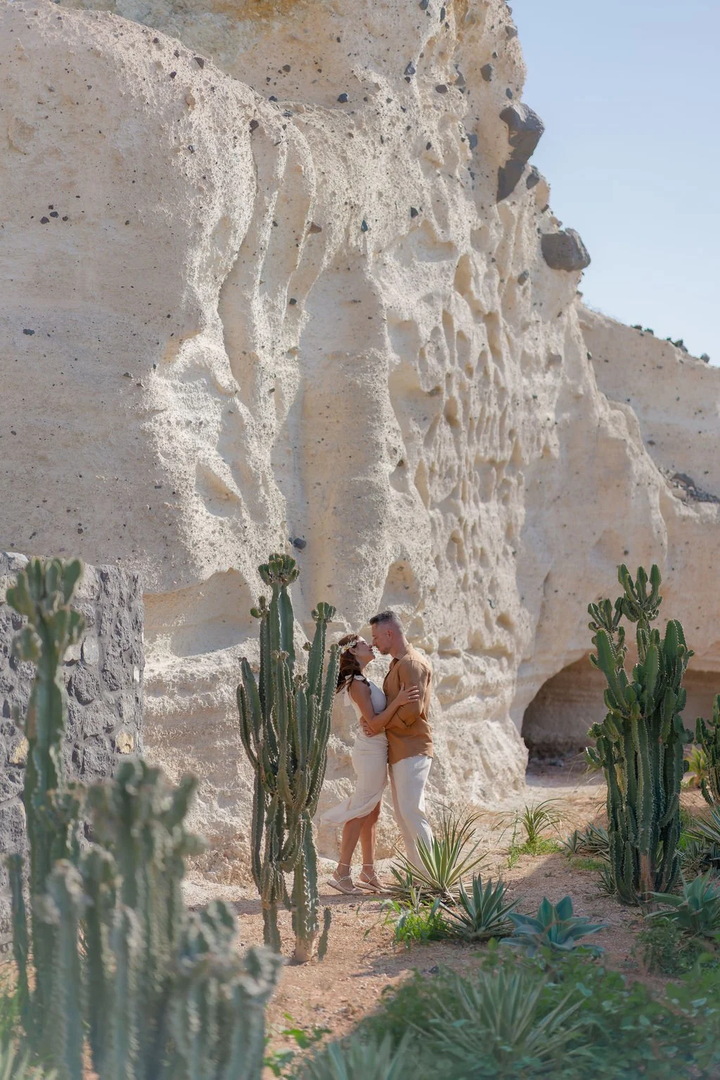 A couple embracing and about to kiss in front of a large, white rocky cliff with cacti and desert plants in the foreground. Proposal preparation in Santorini