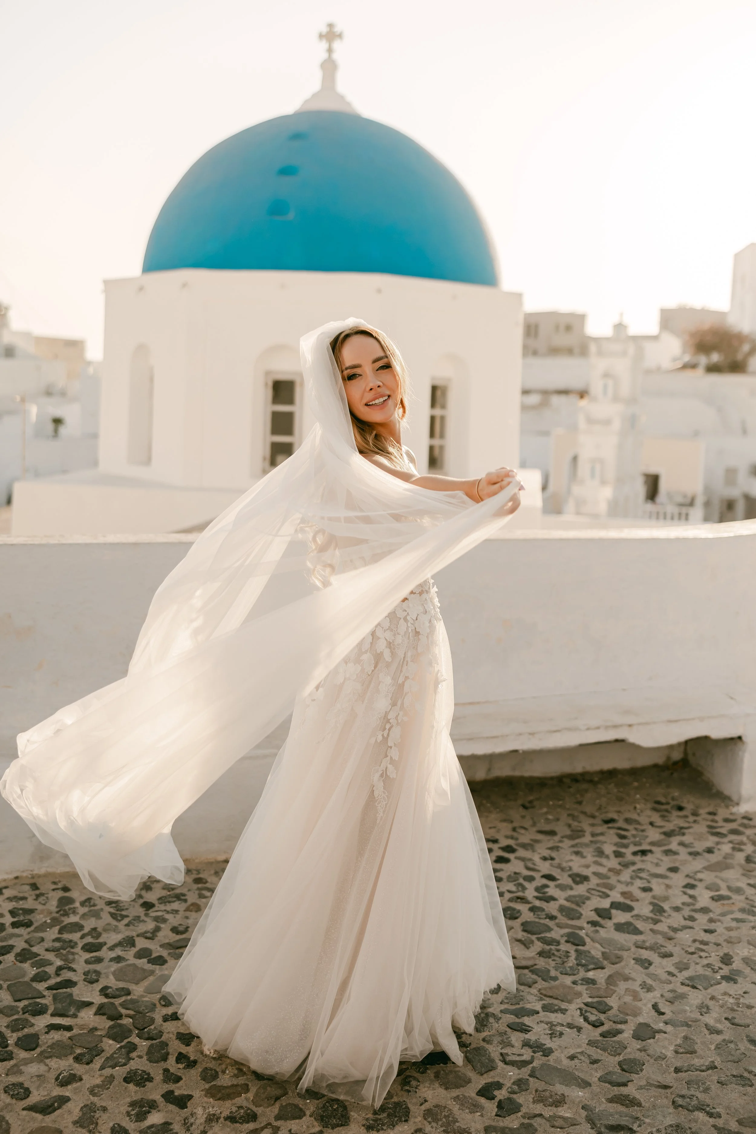 Bride in white wedding dress with veil standing on cobblestone terrace with iconic white buildings and blue-domed church in Santorini, Greece, during sunset.