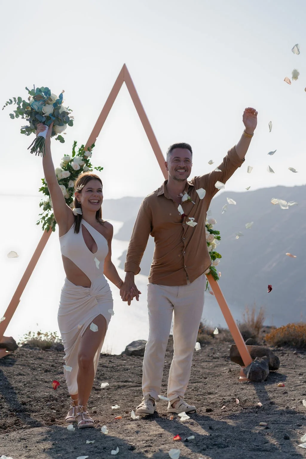 A happy couple holding hands during their wedding celebration outdoors, with a triangular floral arch behind them and flower petals falling around. Just married in Santorini!
