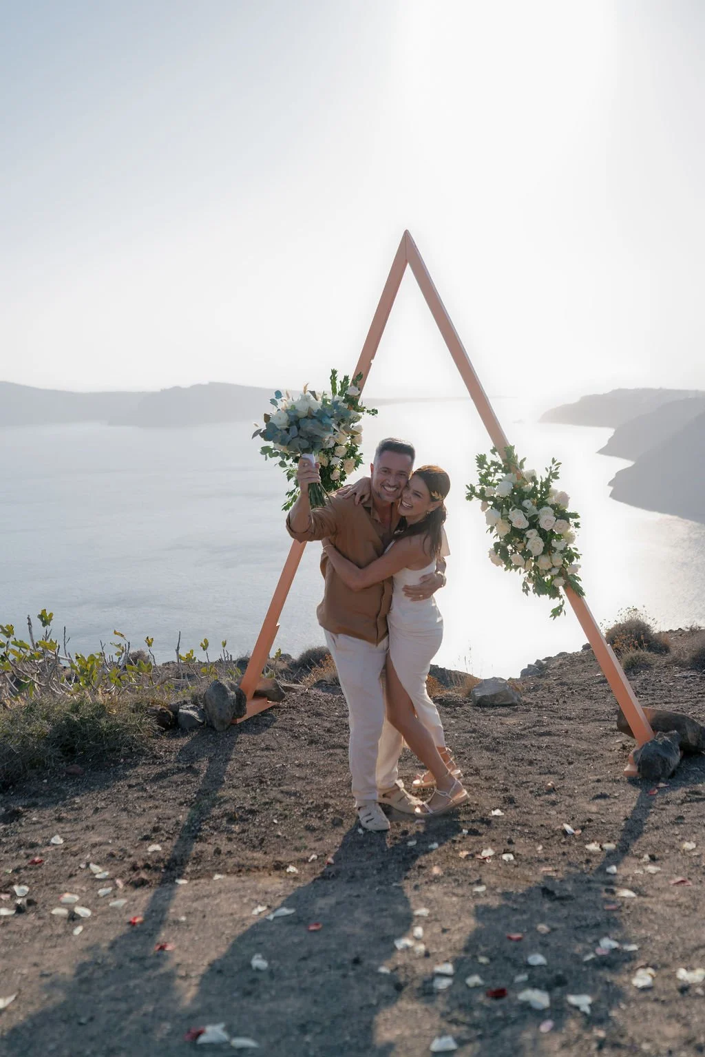 A couple stands happily embracing under a triangular wooden arch decorated with white flowers, overlooking the Santorini caldera at sunset, with scattered rose petals on the ground.