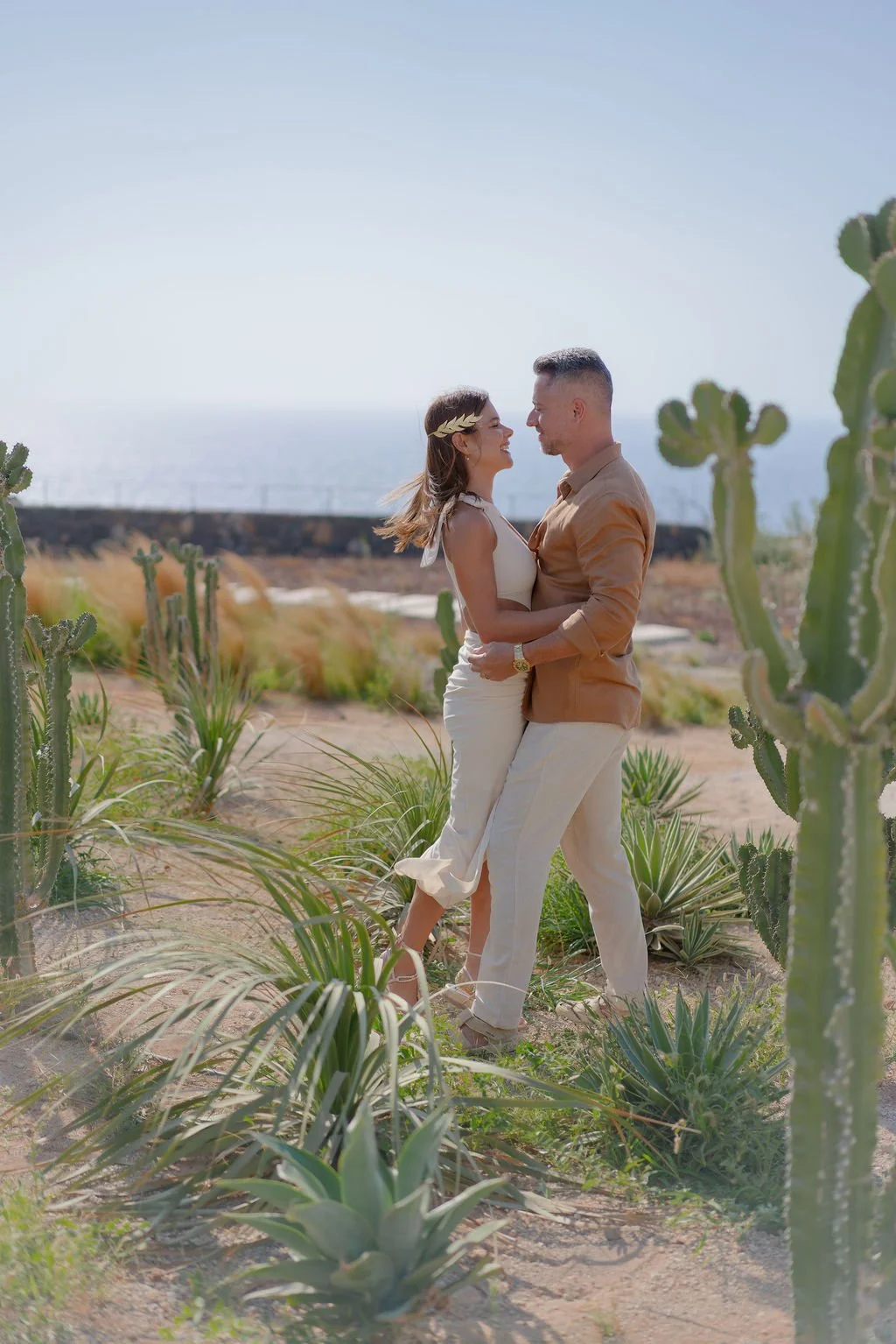 A couple embracing in a desert landscape with cactus plants, overlooking the ocean. Vow renewal in Santorini