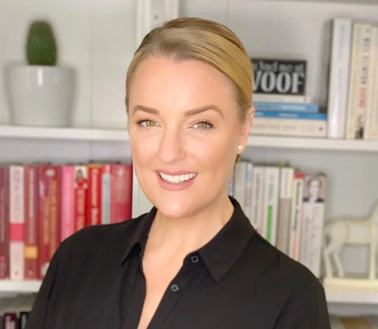 A woman with blonde hair, wearing a black shirt, smiling in front of bookshelves with colorful books and a small potted cactus plant.