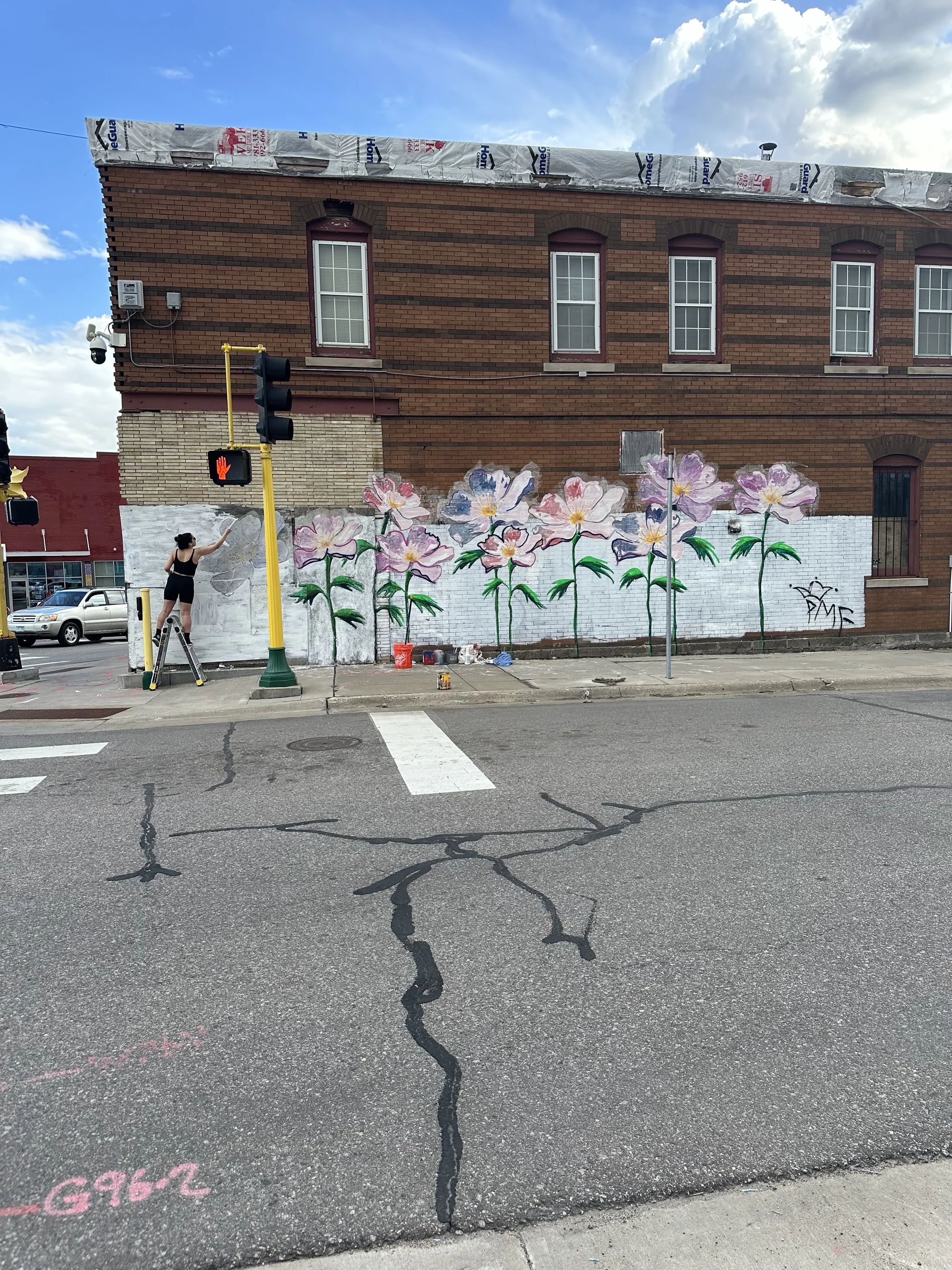A woman on scaffolding painting a mural of large pink, purple, and white flowers on the white lower wall of a brick building. The mural depicts a row of flowers with green stems and petals in various shades of pink, purple, and white.