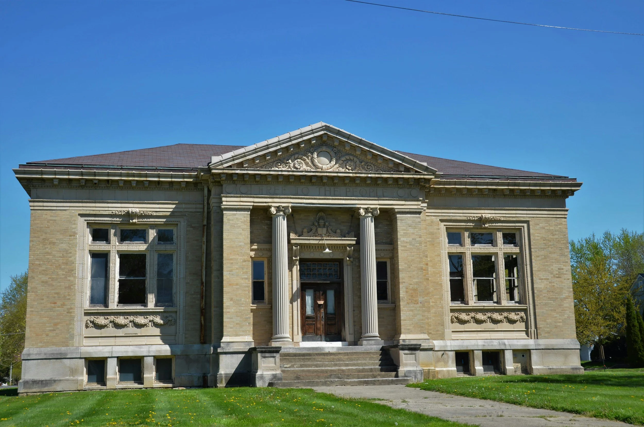 A historic brick building with classical architectural features, including tall columns, decorative moldings, and a triangular pediment, set on a well-maintained lawn with a clear blue sky in the background.