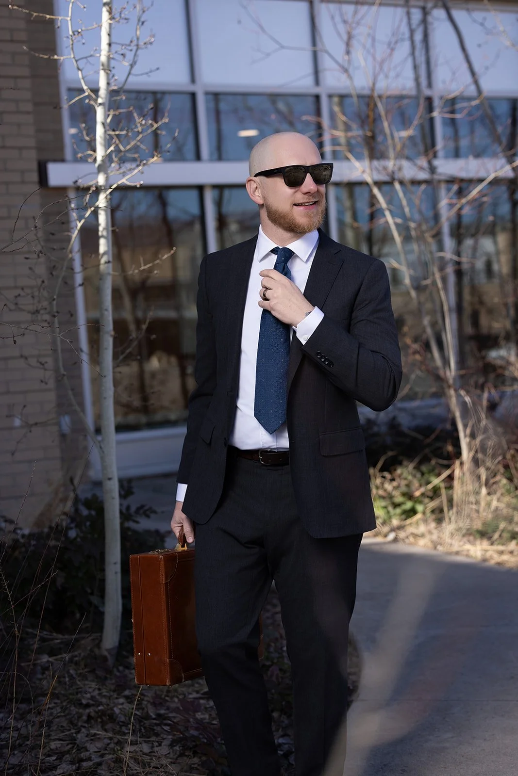 An attorney a beard wearing a black suit, white shirt, and tie, holding a brown briefcase, walking outdoors near the Routt County courthouse with glass windows and leafless trees, wearing sunglasses.