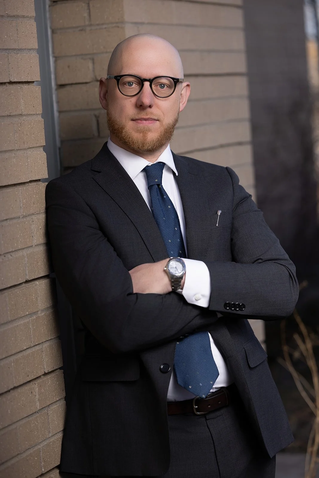 A professional man with glasses and a beard, wearing a dark suit, white shirt, and blue tie, standing with arms crossed against a brick wall.
