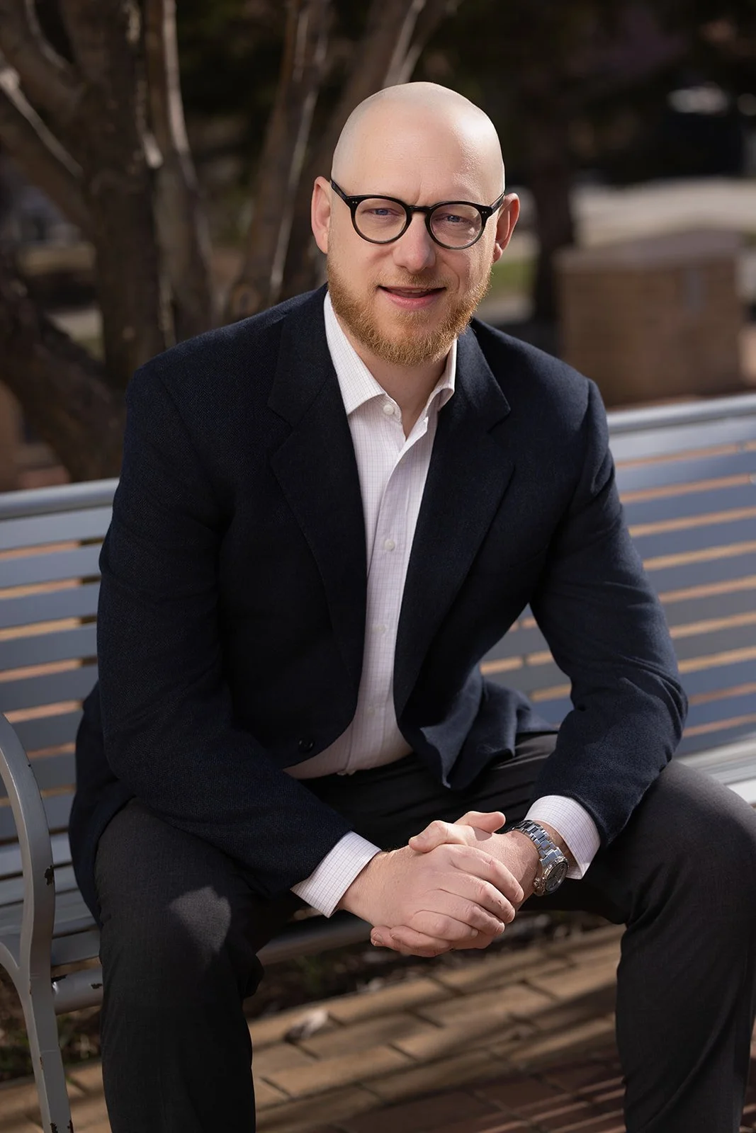 A smiling man with glasses, a bald head, and a beard, sitting on a park bench outdoors, wearing a dark blazer, white shirt, and black pants.