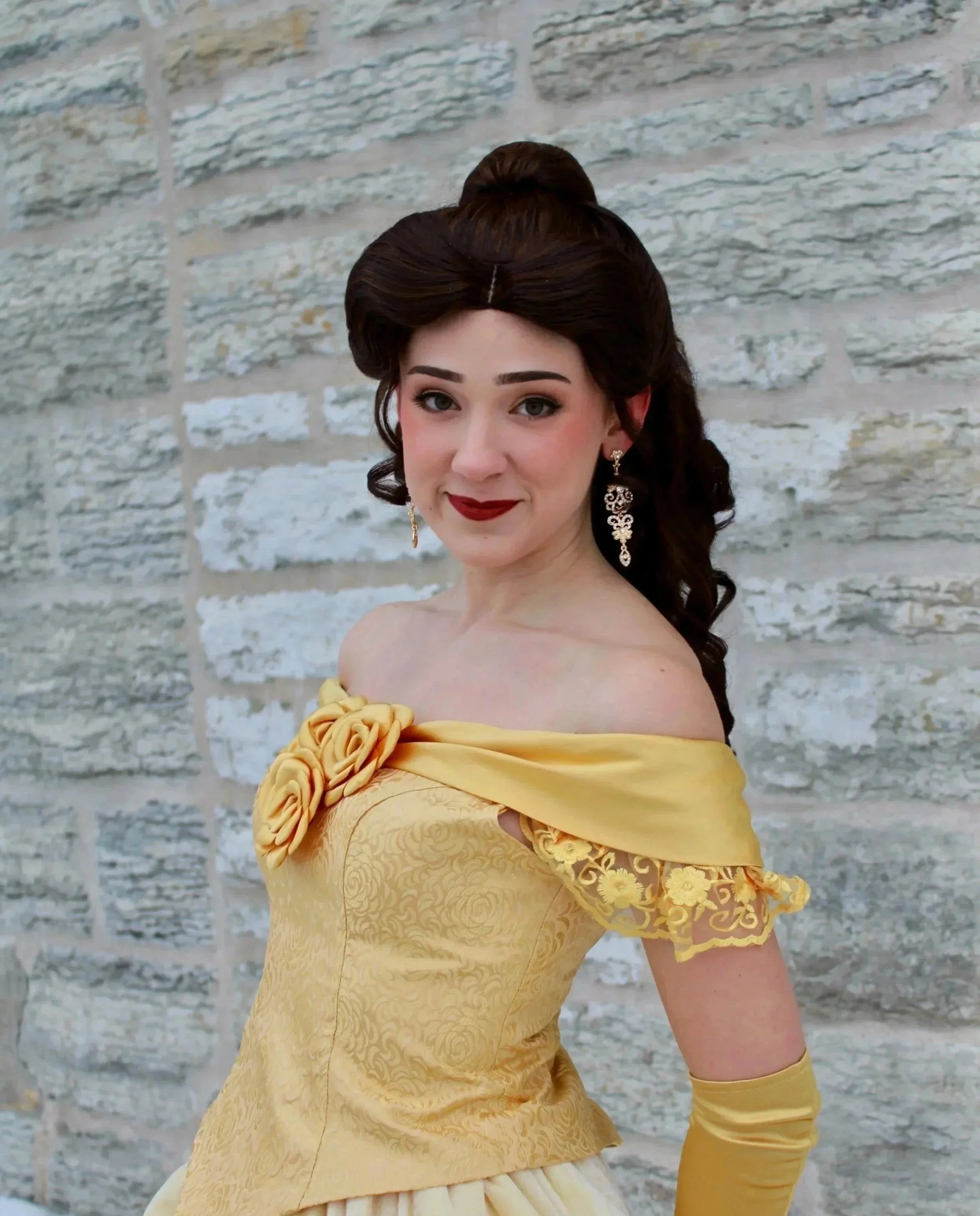 Princess Belle wearing a yellow off-the-shoulder gown with floral embellishments, standing in front of a stone wall, wearing long earrings and red lipstick.