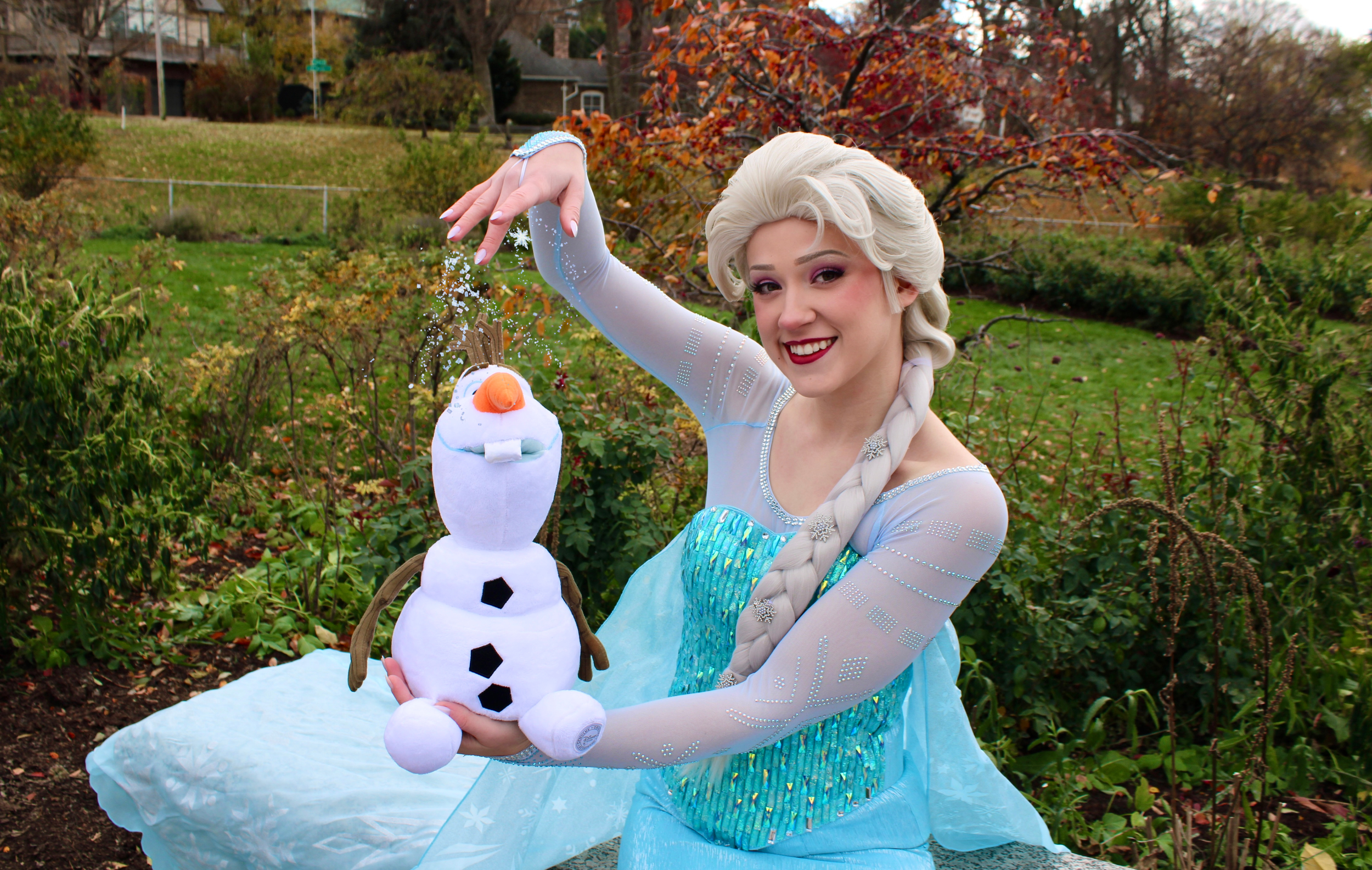 Elsa holding a plush snowman toy, standing outdoors in a garden with fall foliage in the background.