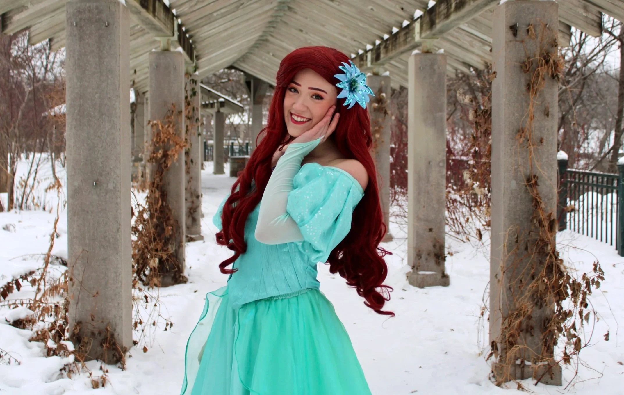 Ariel with long red hair wearing an off-the-shoulder aqua dress and a large blue flower in her hair, standing under a covered walkway in a snowy outdoor setting, smiling and posing with her hand on her cheek.
