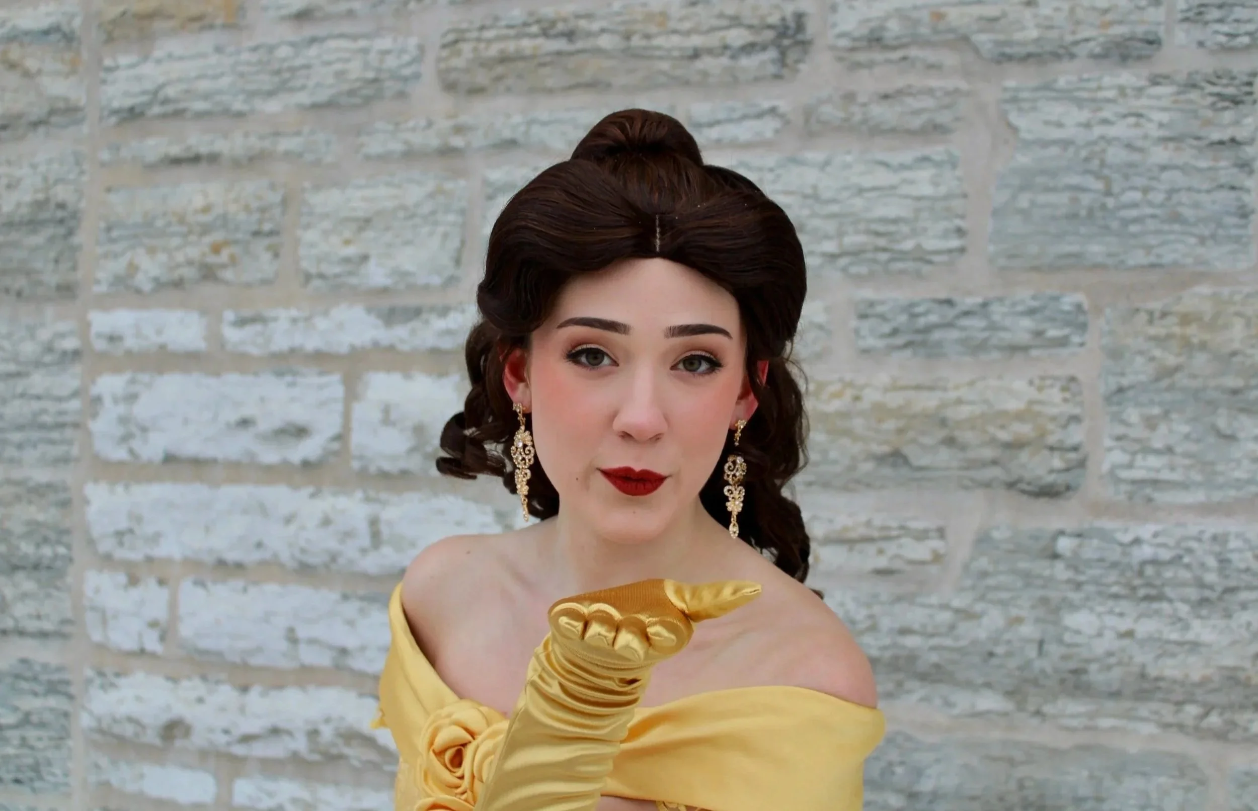 Princess Belle wearing a yellow gown with long gloves, pointing at the camera, standing against a light-colored brick wall.