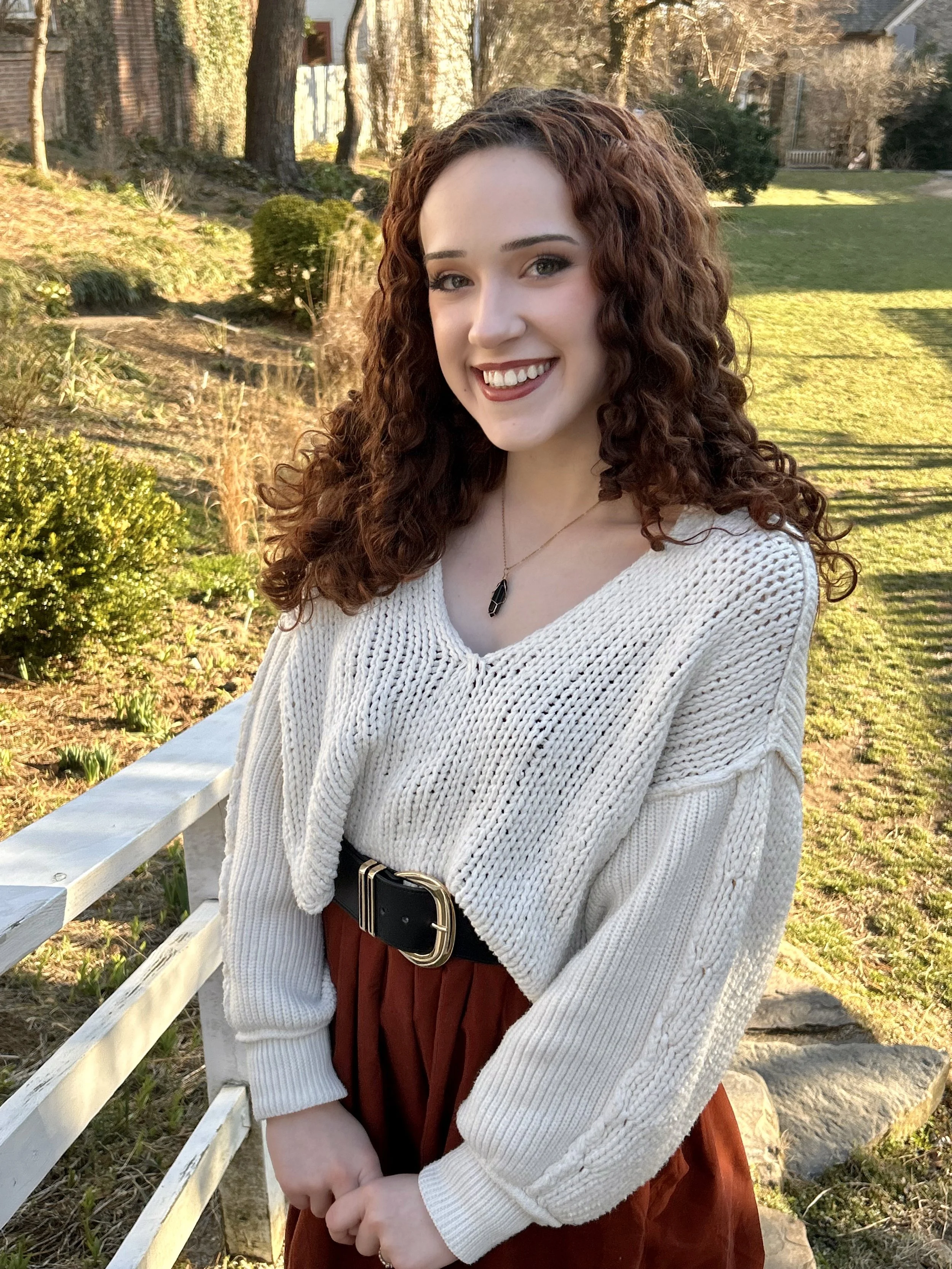 A young woman with curly red hair and fair skin smiling outdoors in a garden during late afternoon, wearing a white knitted sweater, a black belt, a burgundy skirt, and a necklace with a black pendant.