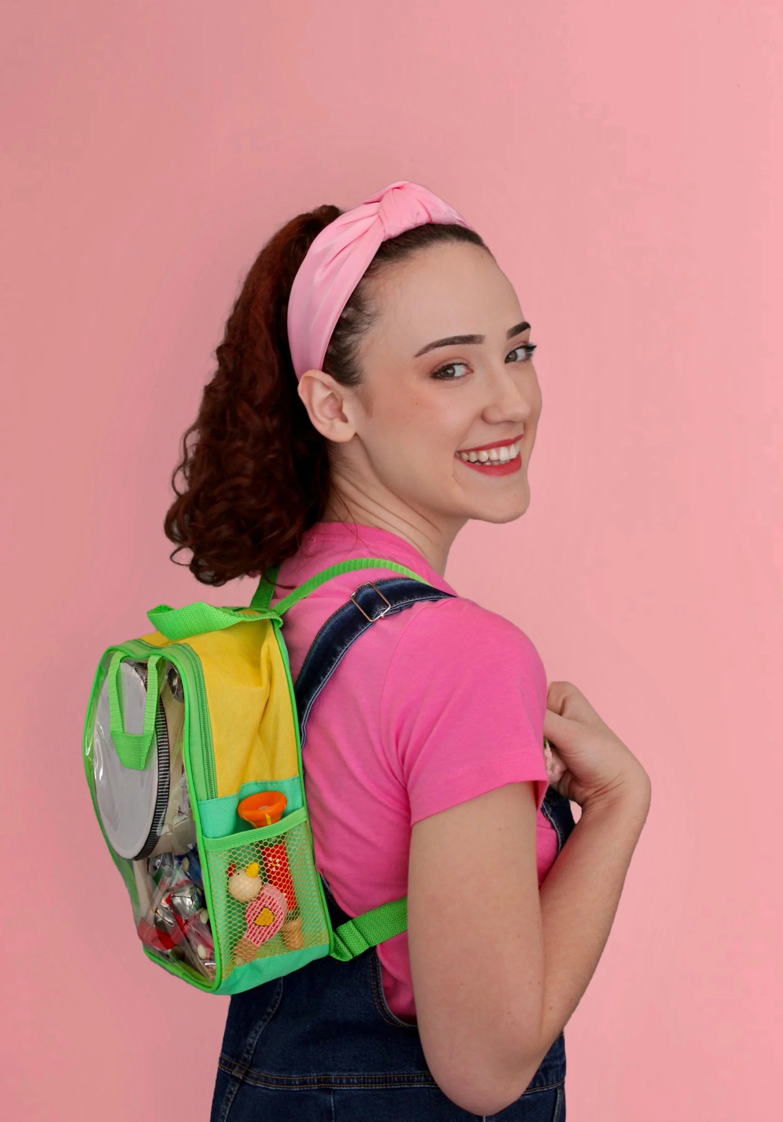 Miss Rach wearing a pink headband, pink t-shirt, and denim overalls, carrying a colorful backpack, smiling at the camera, standing against a pink background.