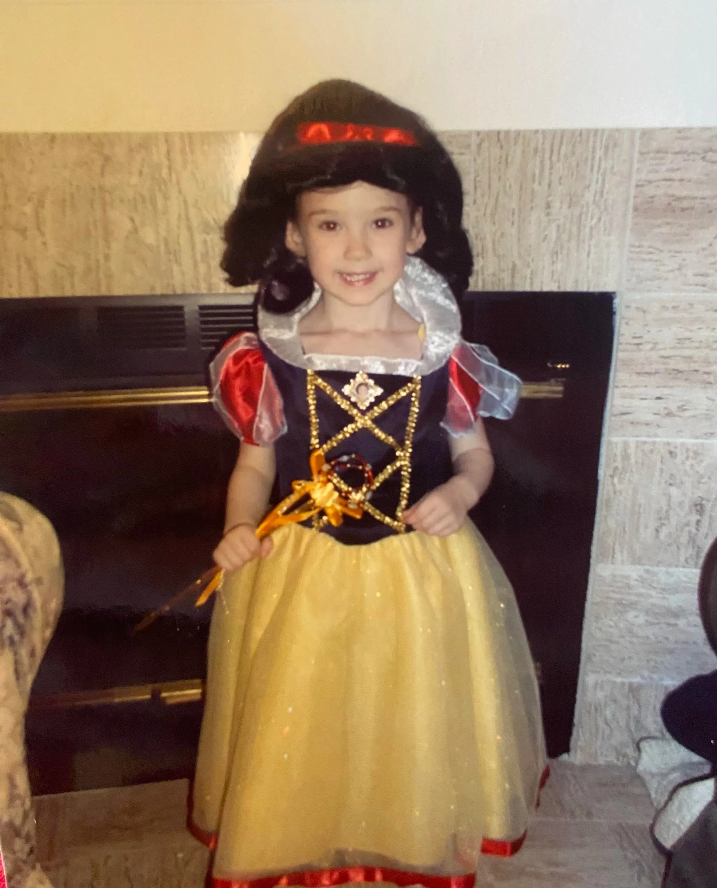 A young girl dressed as Snow White, wearing a costume with a yellow skirt, blue and red top, and a black wig with a red bow, standing indoors in front of a fireplace mantle.