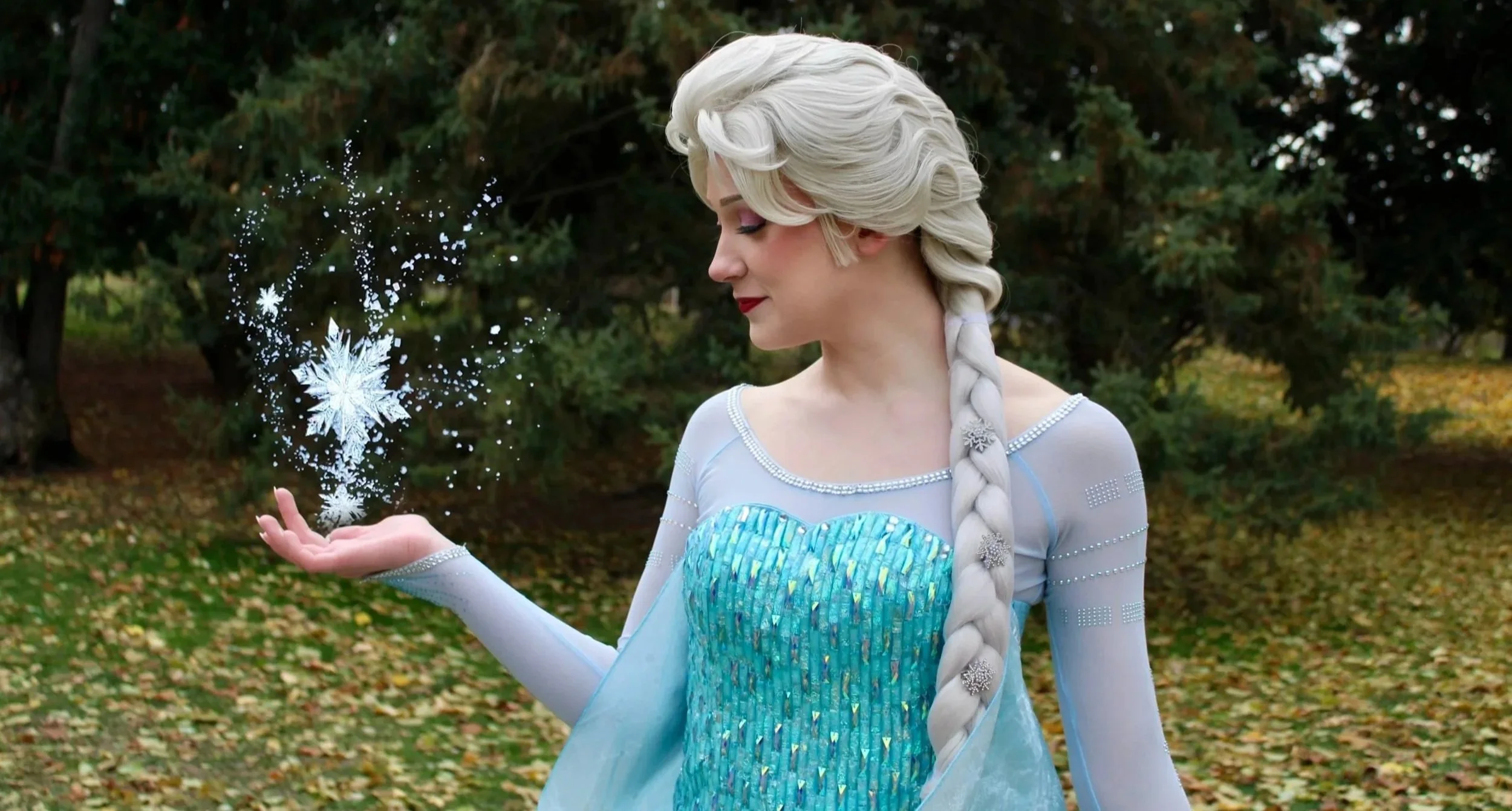 Elsa from Frozen standing in a park with trees and fallen leaves, holding glittering snowflake magic in her hand.