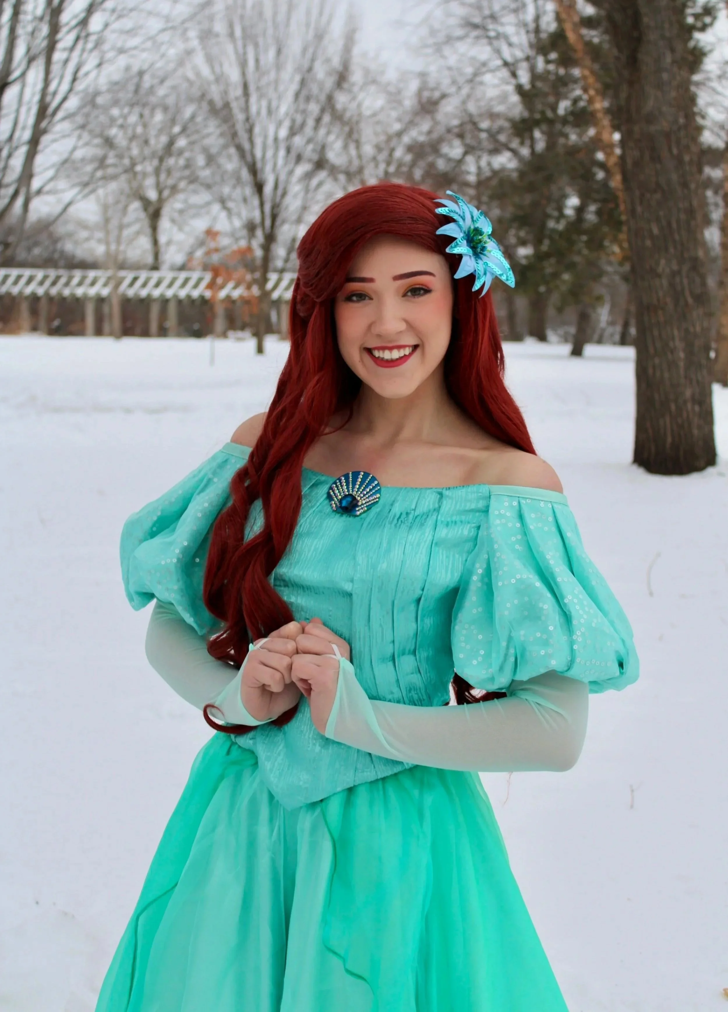 Ariel in a turquoise dress, with long red hair and a blue flower hair accessory, standing outdoors in a snowy park with bare trees.