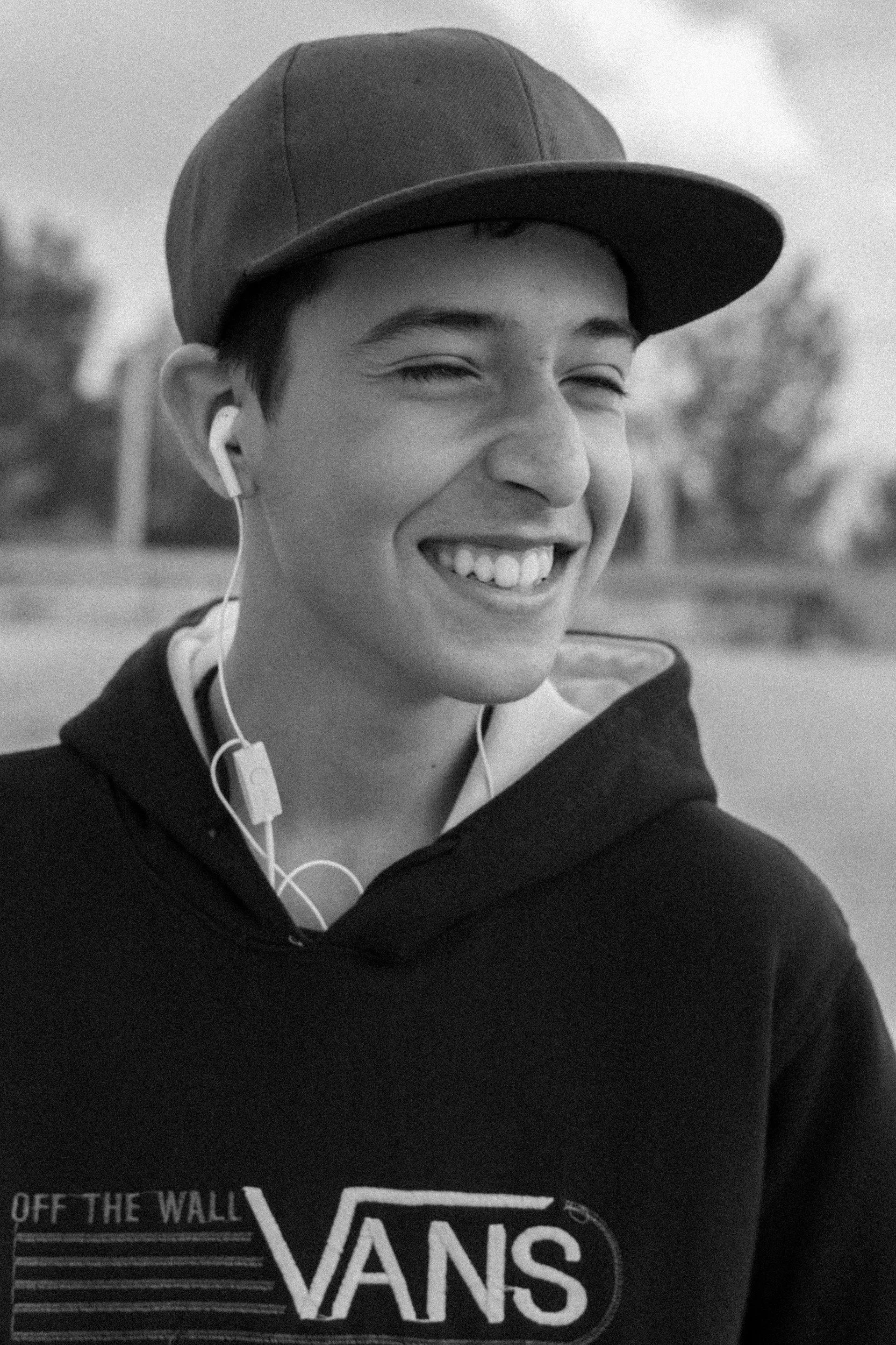 A smiling young man wearing a baseball cap, hoodie with Vans logo, and wireless earbuds outdoors.
