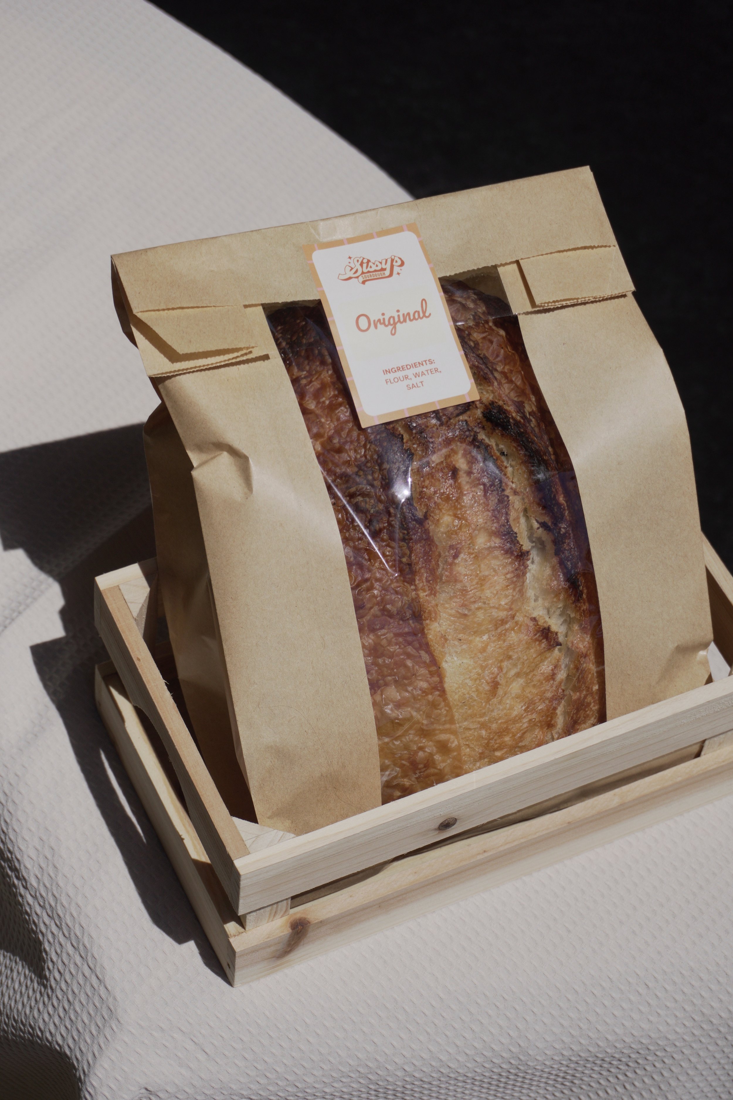 A loaf of bread in a brown paper bag with a clear window, labeled "Original," sitting in a small wooden crate on a white textured surface.