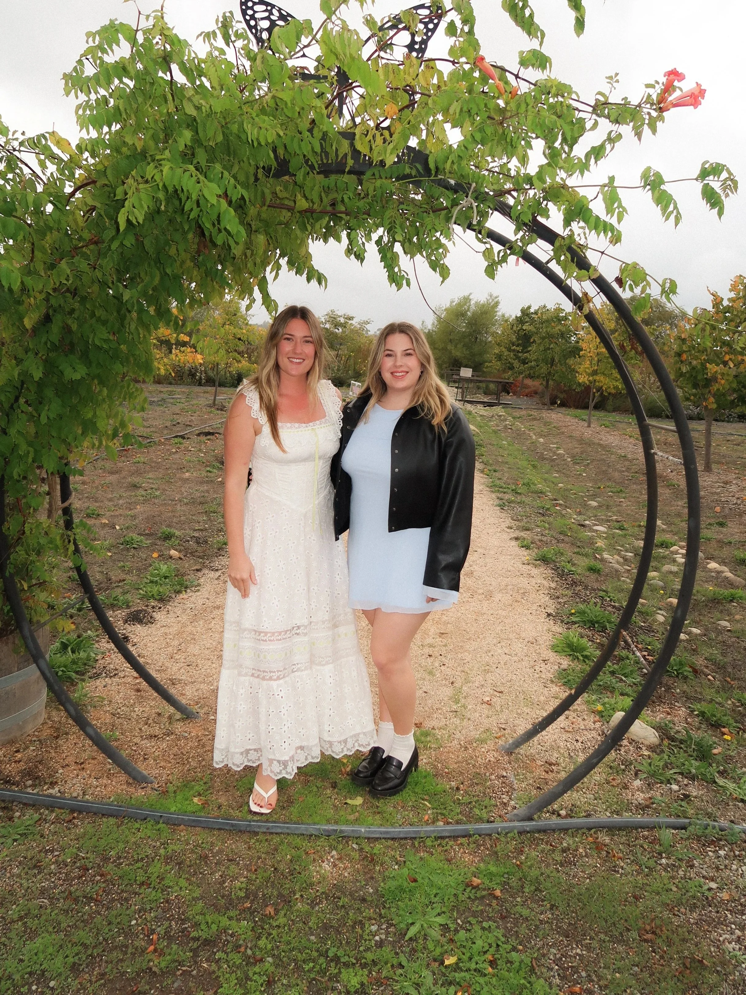 Two women standing outdoors in a vineyard or garden, posing for a photo inside a circular metal arch with green foliage and pink flowers above them. One woman is wearing a white lace dress, and the other is in a light blue dress with a black leather jacket. They are smiling at the camera.