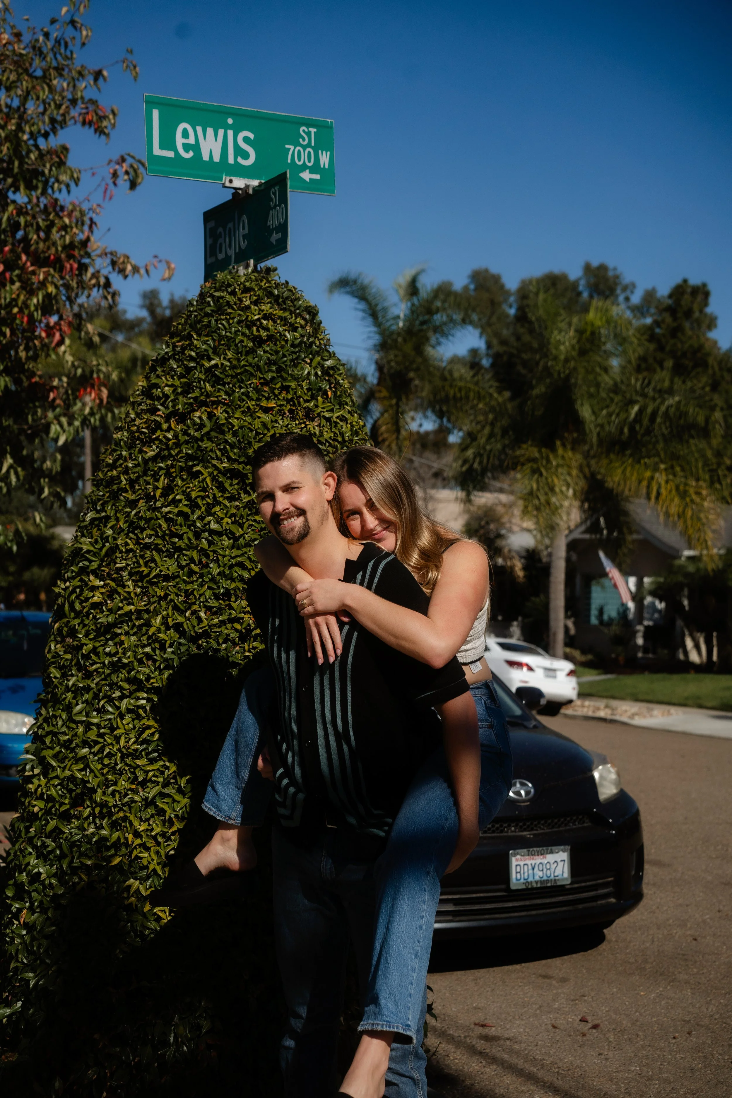 A happy couple with a man giving a piggyback ride to a woman, posing near street signs that read Lewis Street and Eagle Street, over a background of trees, cars, and a neighborhood under a clear blue sky.
