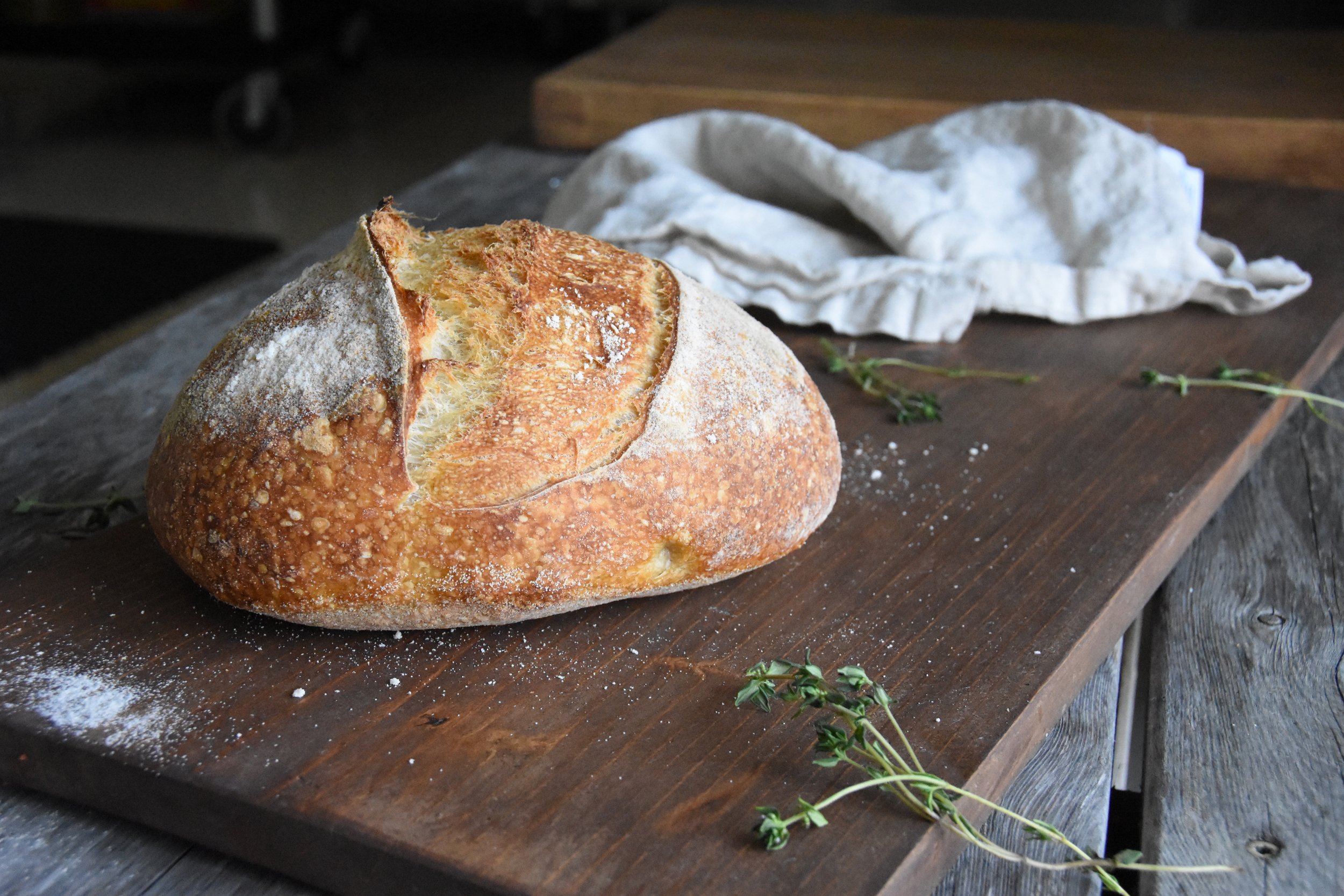 A rustic loaf of bread with a golden-brown crust and a cracked top sits on a wooden cutting board. A white cloth and a sprig of thyme rest nearby on the wooden surface.
