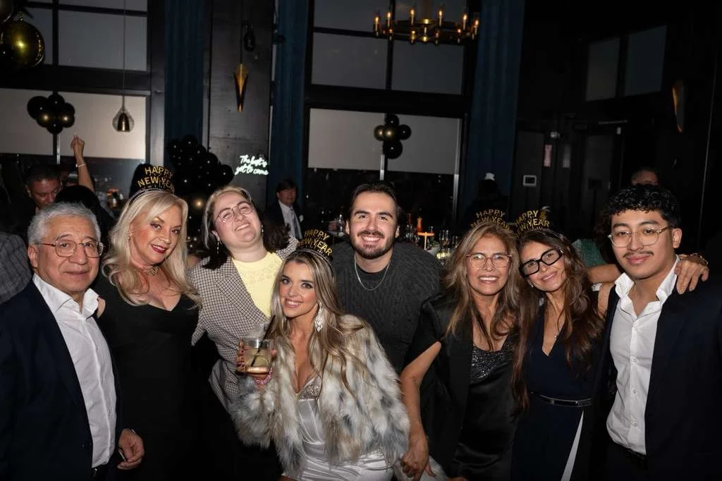 Group of people celebrating New Year's Eve at a party in a decorated indoor venue, wearing 'Happy New Year' headbands, smiling, with champagne glasses and decorations in the background.