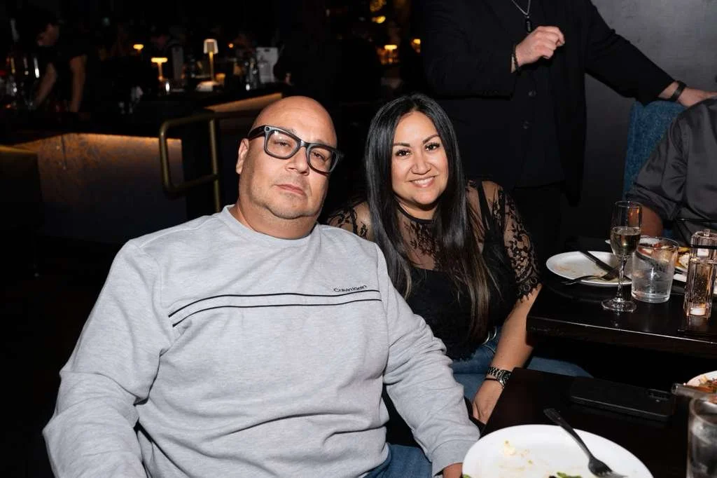 A man and woman sitting together at a restaurant table, smiling, with drinks and plates on the table, in a dimly lit, upscale dining setting.
