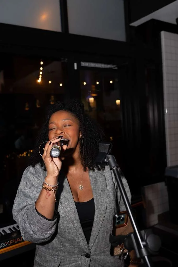A woman singing into a microphone in a dimly lit room, likely a music venue or bar, standing near a keyboard and a music stand.