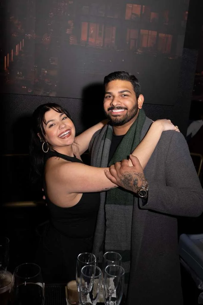 Smiling woman and man embracing at a social event, with glasses on the table in front of them.