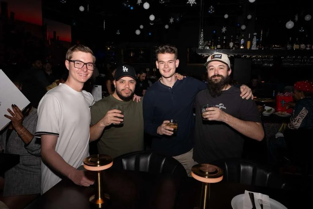 Four men standing together at a bar, holding drinks, smiling at the camera, with a dark background decorated with hanging snowflakes and ornaments.