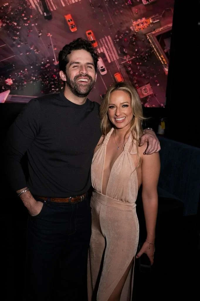 A man and woman smiling and posing together indoors, with a cityscape backdrop projected on a screen behind them.