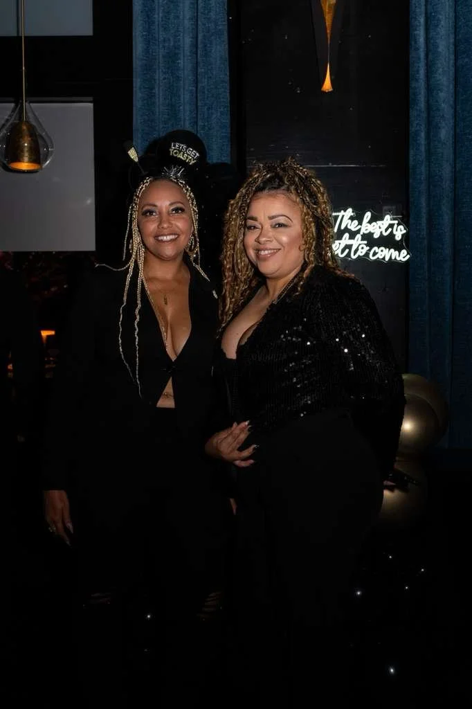 Two women dressed in black, smiling, at a celebration or party with dark blue curtains, a neon sign, and black and gold balloons in the background.