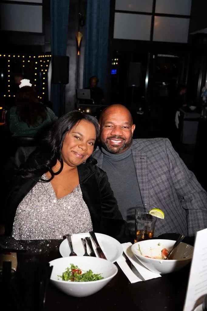 A couple sitting at a restaurant table, smiling at the camera. The woman is wearing a sparkly silver top and black jacket, and the man is in a gray plaid blazer and turtleneck. The table has salad, bowls, utensils, a glass with a lemon slice, and the