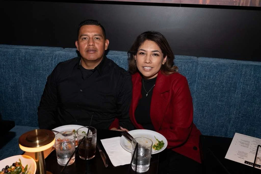 A man and woman sitting at a restaurant table with plates of food and drinks, smiling at the camera.