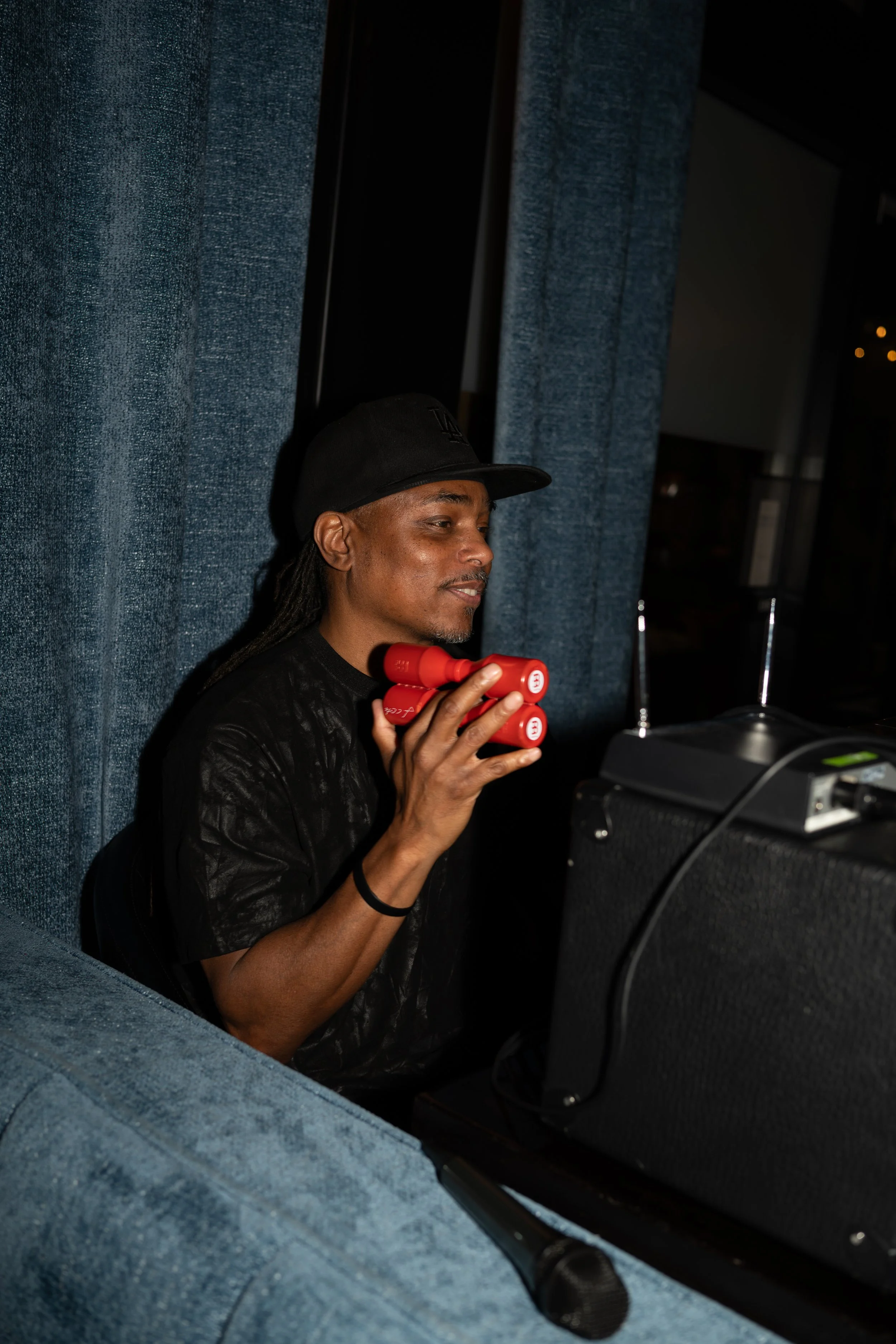 A man sitting on the floor, wearing a black hat and black shirt, holds three red cylindrical objects in his hand, with a black speaker and electronic equipment nearby. He is partially framed by a blue fabric surface.