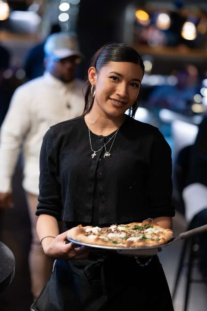 A young woman holding a pizza on a plate, smiling, in a dimly lit restaurant or pizzeria. She has dark hair, wears a black shirt, and has multiple necklaces.